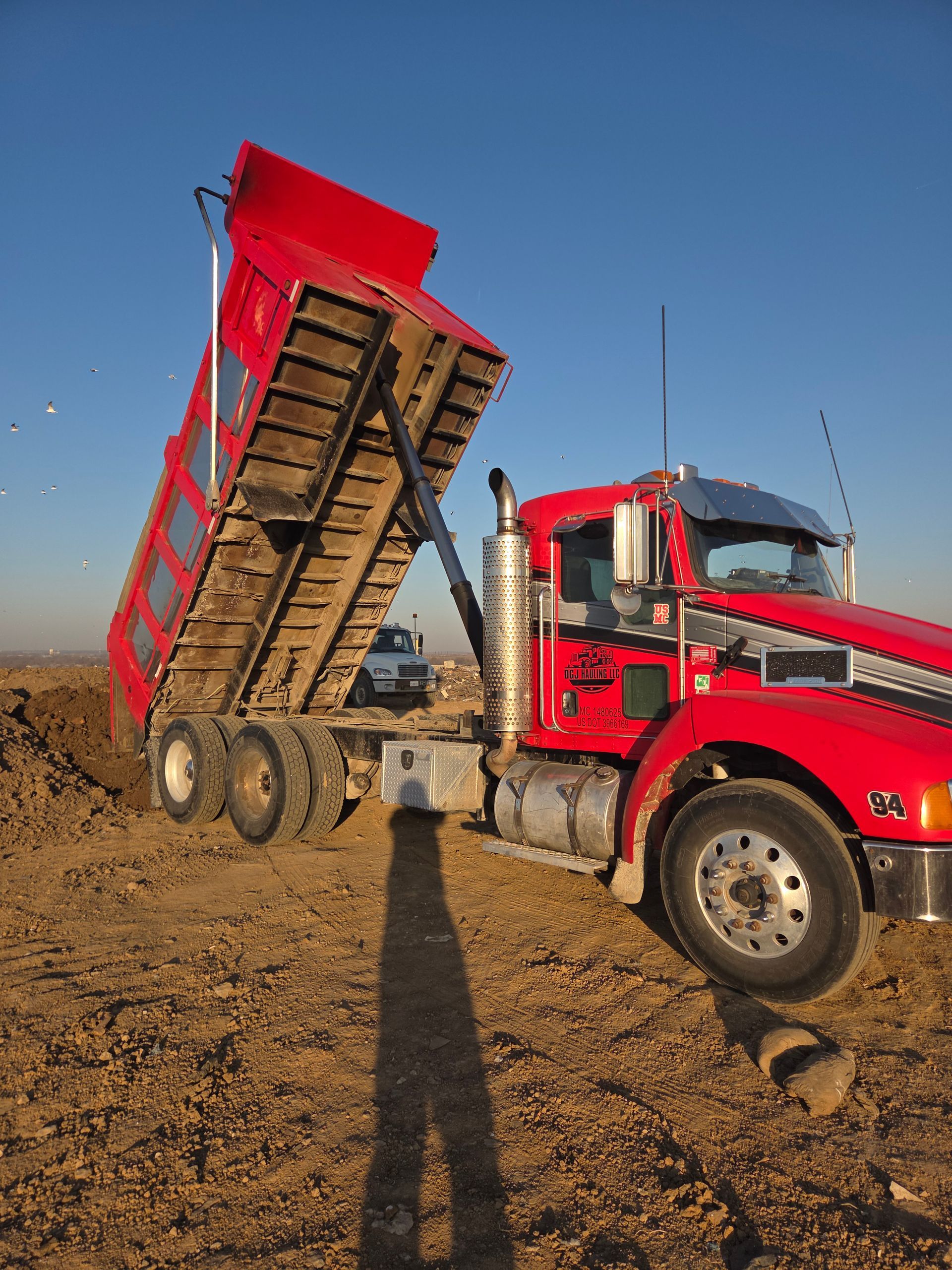 Red dump truck with raised bed unloading on a dirt field under a blue sky.