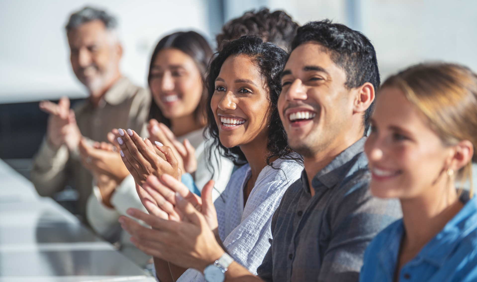 A row of diverse employees sit at an office table, clapping in recognition and praise of coworkers