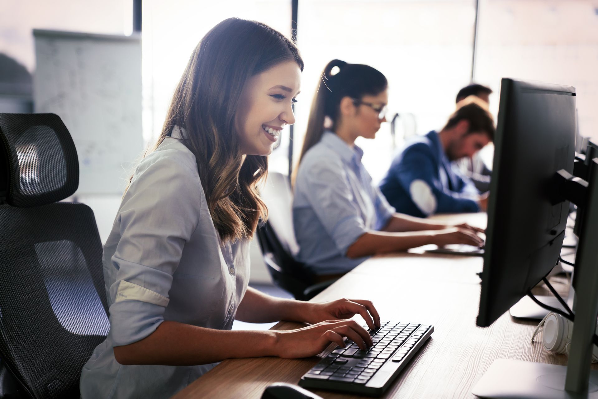 White woman smiles at her computer while working, coworkers behind her in the distance work on their computers