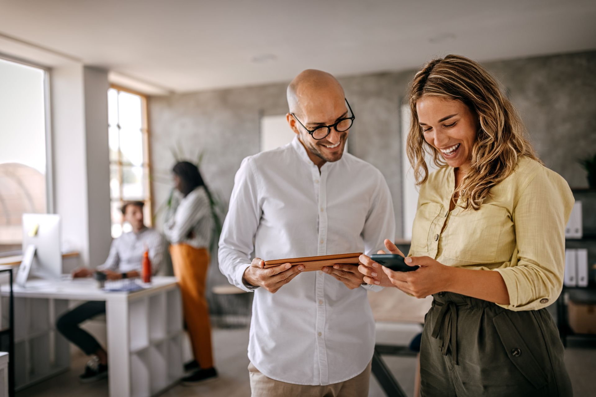 Coworkers, a white man with glasses and a white woman, stand each holding a tablet