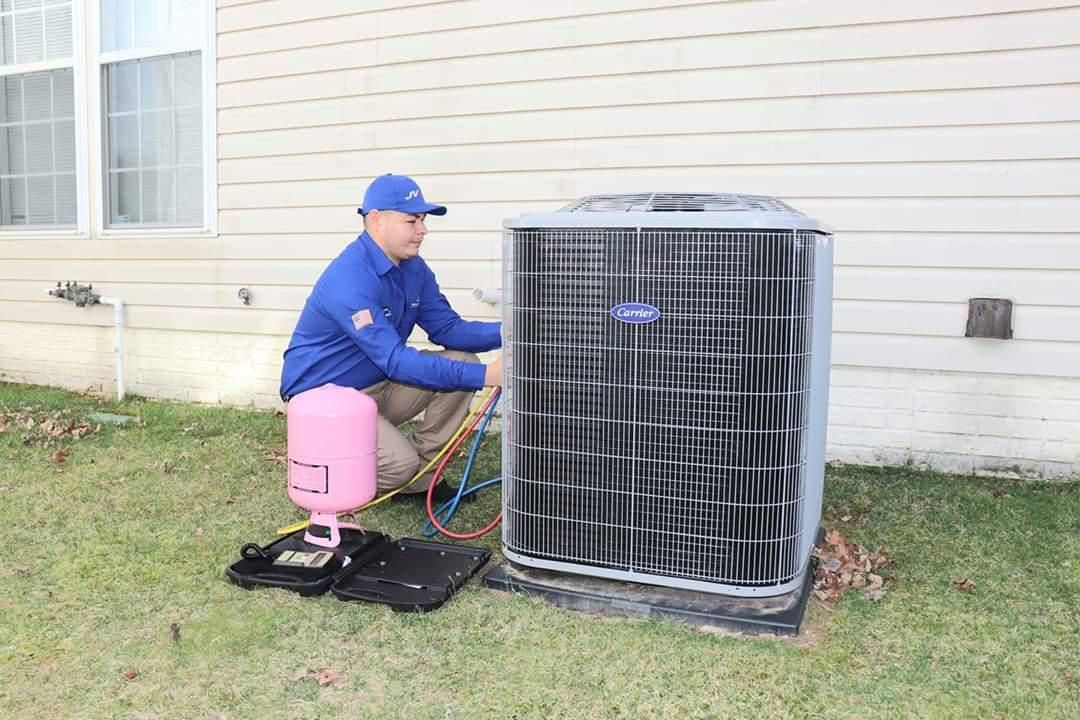 A technician in a blue uniform services an outdoor air conditioning unit with a refrigerant cylinder on the grass.