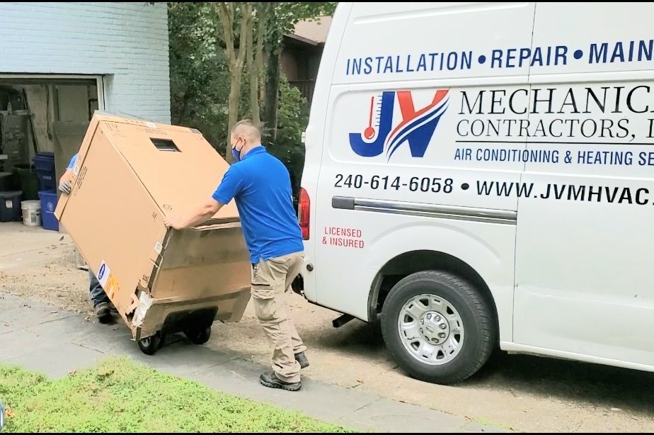 A technician in a blue shirt pushes a boxed HVAC unit toward a JV Mechanical Contractors van parked in a driveway.