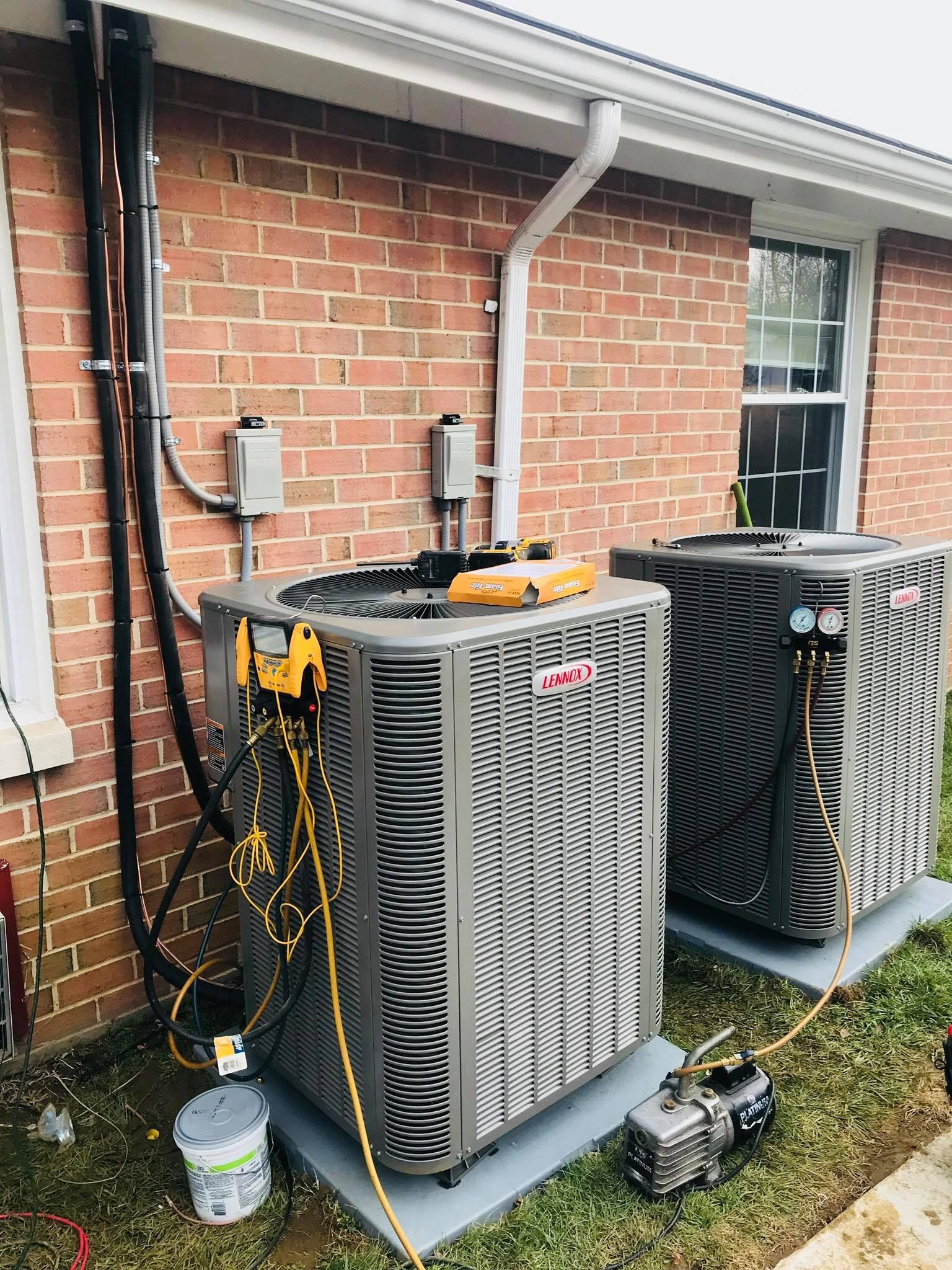 Two gray outdoor HVAC condenser units installed against a brick wall, connected to diagnostic tools and a vacuum pump.