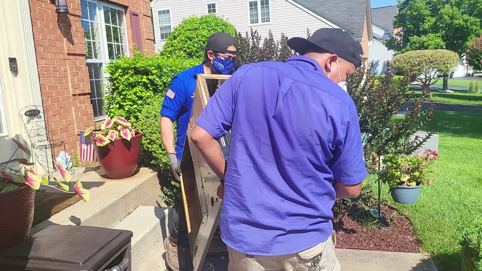 Two people wearing hats and matching blue shirts move a large, flat object outdoors on a residential driveway.