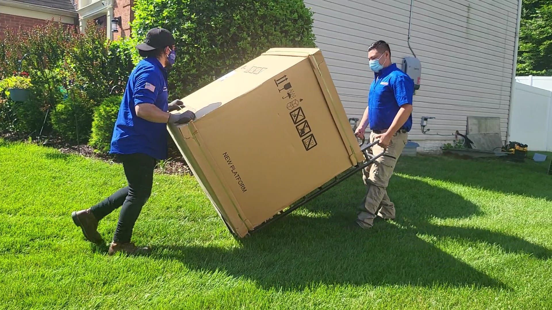 Two people wearing blue shirts and masks move a large, boxed appliance across a lawn using a hand truck.