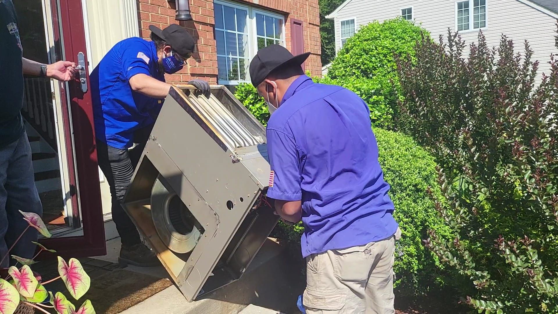 Two workers in blue uniforms lift a metal HVAC air handler unit outside a brick house.