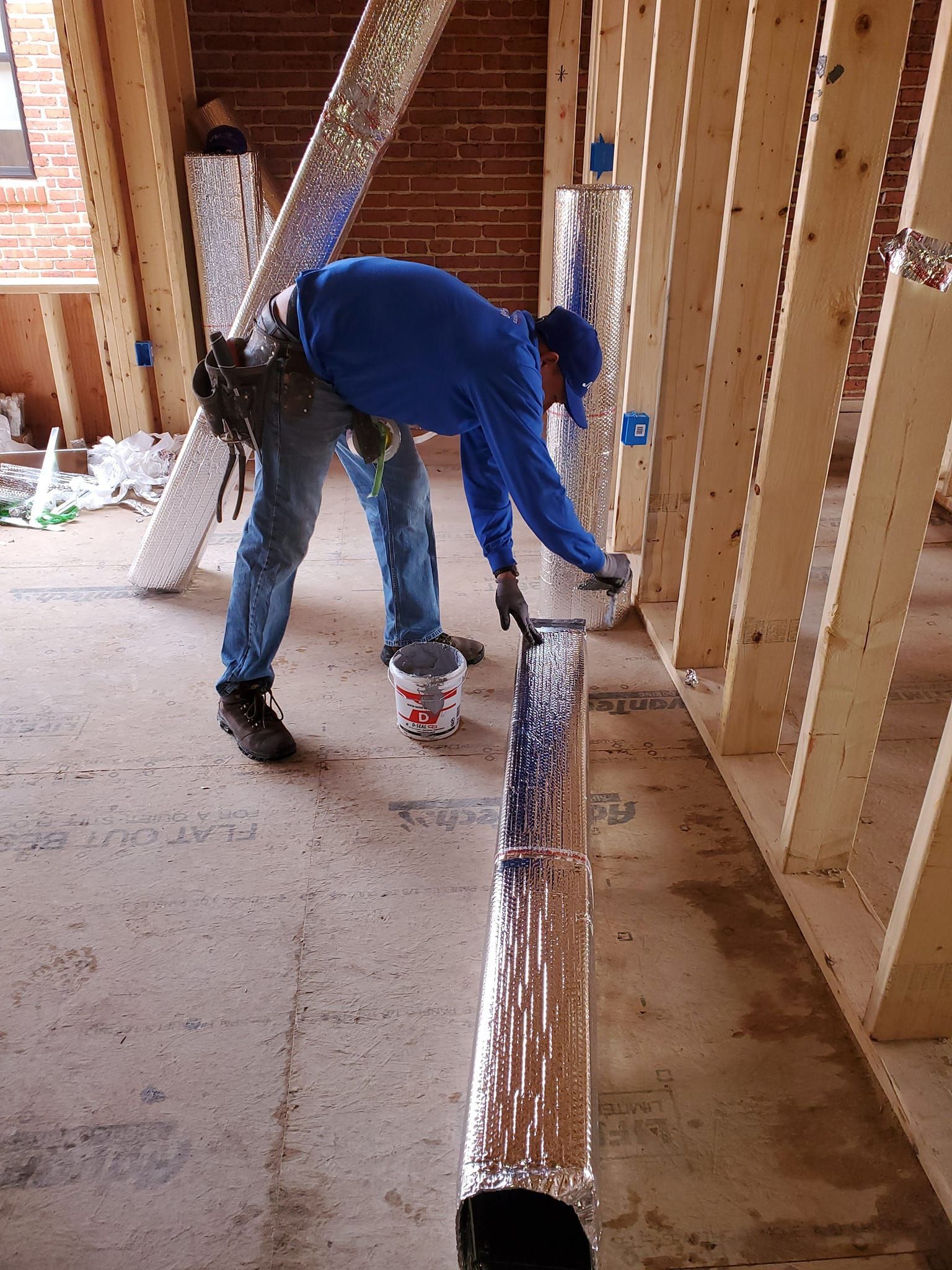 A person in a blue shirt applies foil-backed insulation tape to a rectangular duct inside a wooden-framed building.