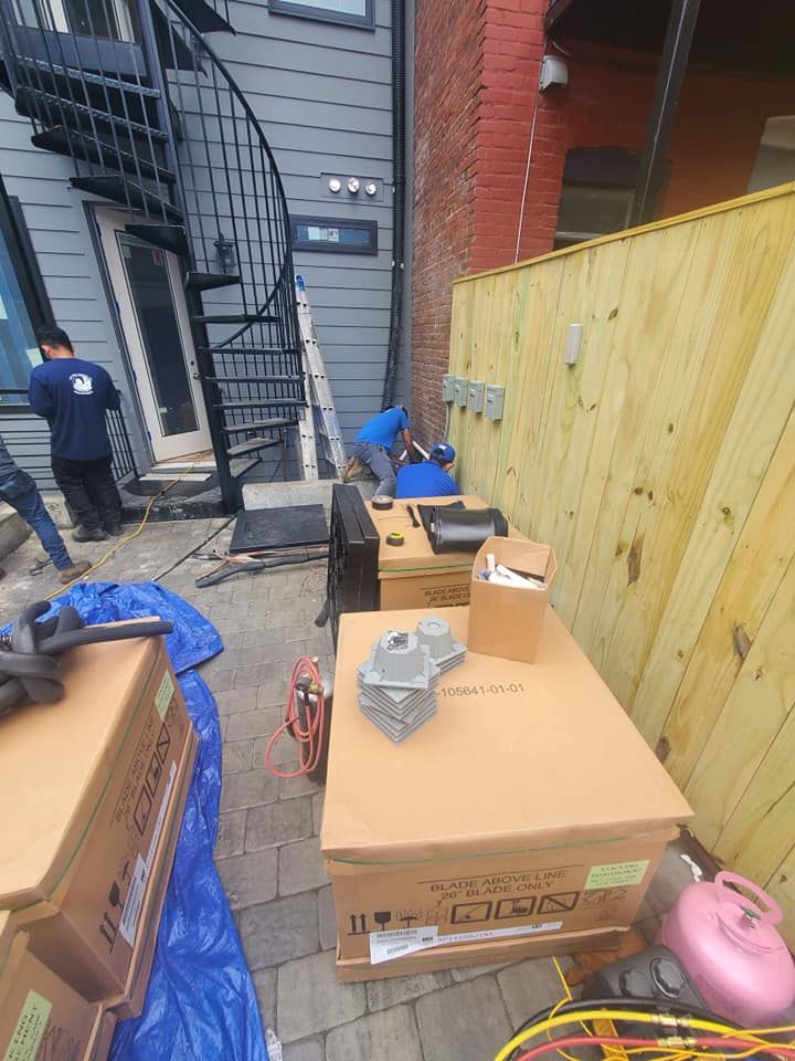 Workers install exterior equipment near a spiral staircase and a wood fence in a paved outdoor space with equipment boxes.