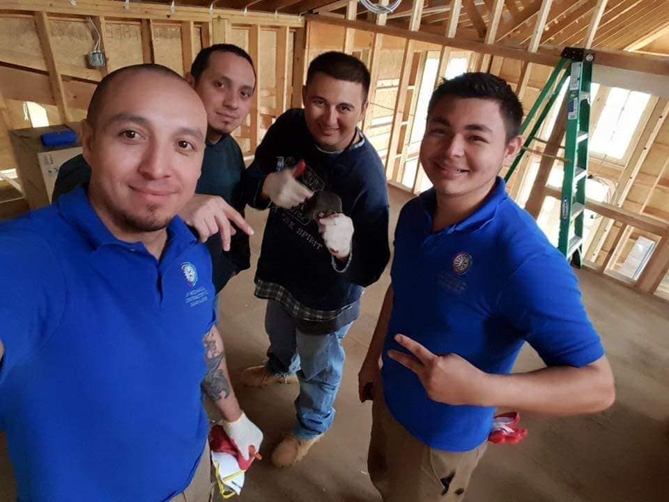 Four people in a construction site interior pose for a selfie, wearing blue shirts and work gear, smiling at the camera.