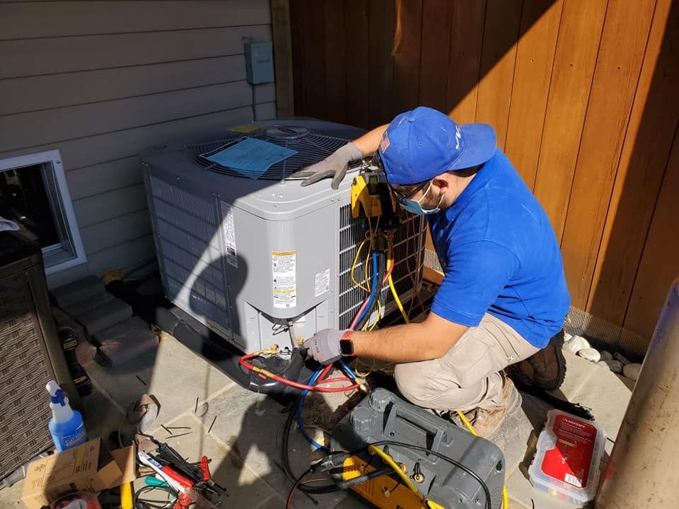 A technician in a blue uniform kneels to repair an outdoor HVAC unit, using tools and equipment on a patio.