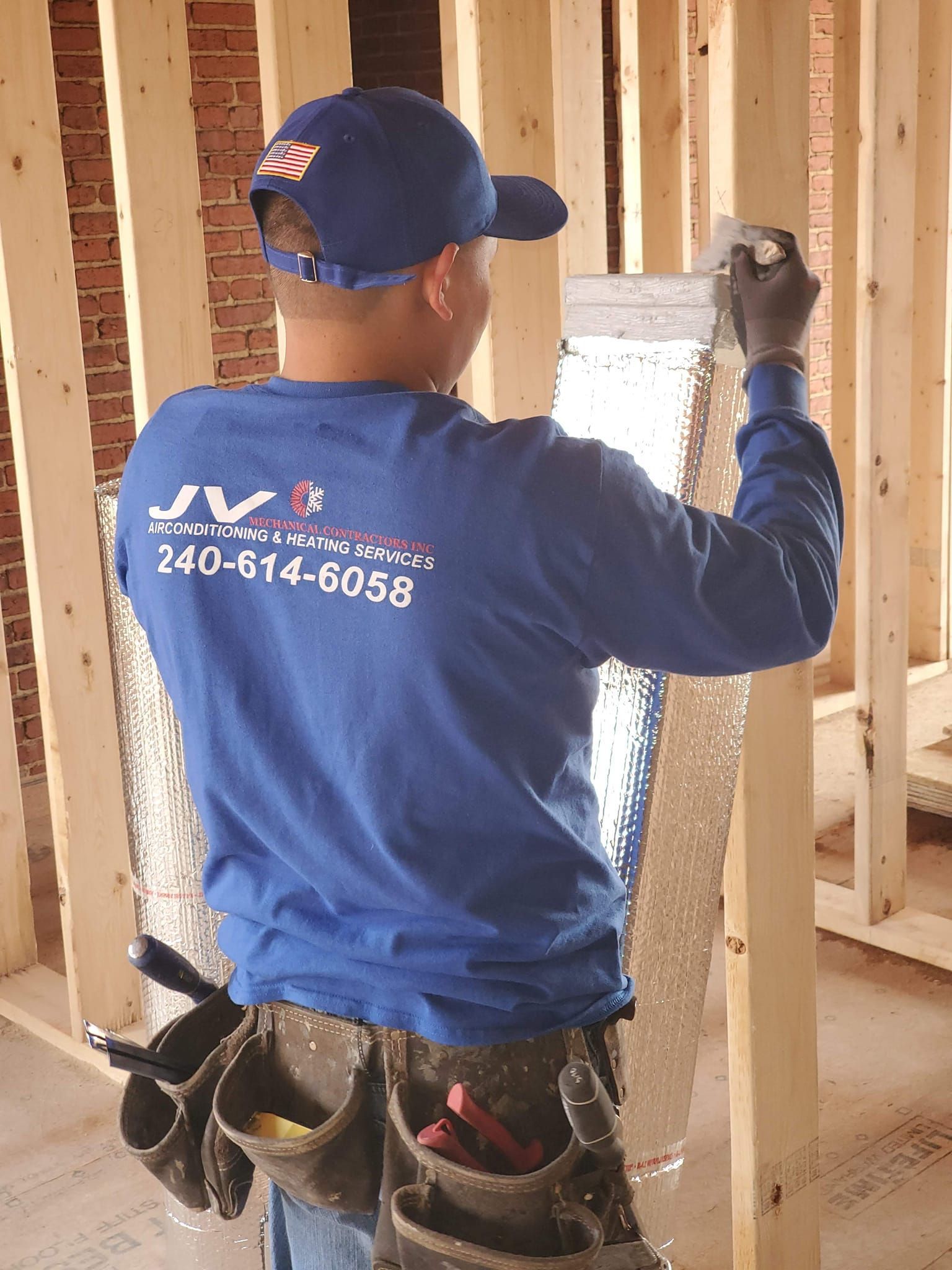 A worker wearing a blue shirt and cap installs metallic reflective insulation between wooden wall studs.