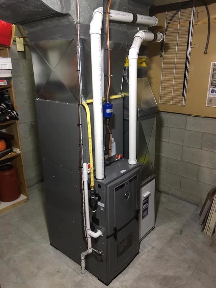 A gray HVAC furnace unit stands in a basement with connected white PVC venting pipes and a yellow gas line.