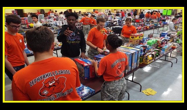 A group of people are standing around a table with toys on it and one of them is wearing an orange shirt that says army