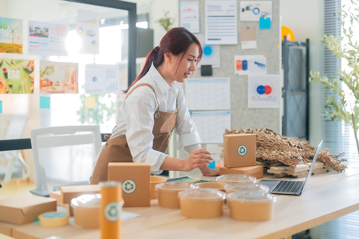 A woman works at her desk filled with packaging materials, focused on her laptop in an office space.