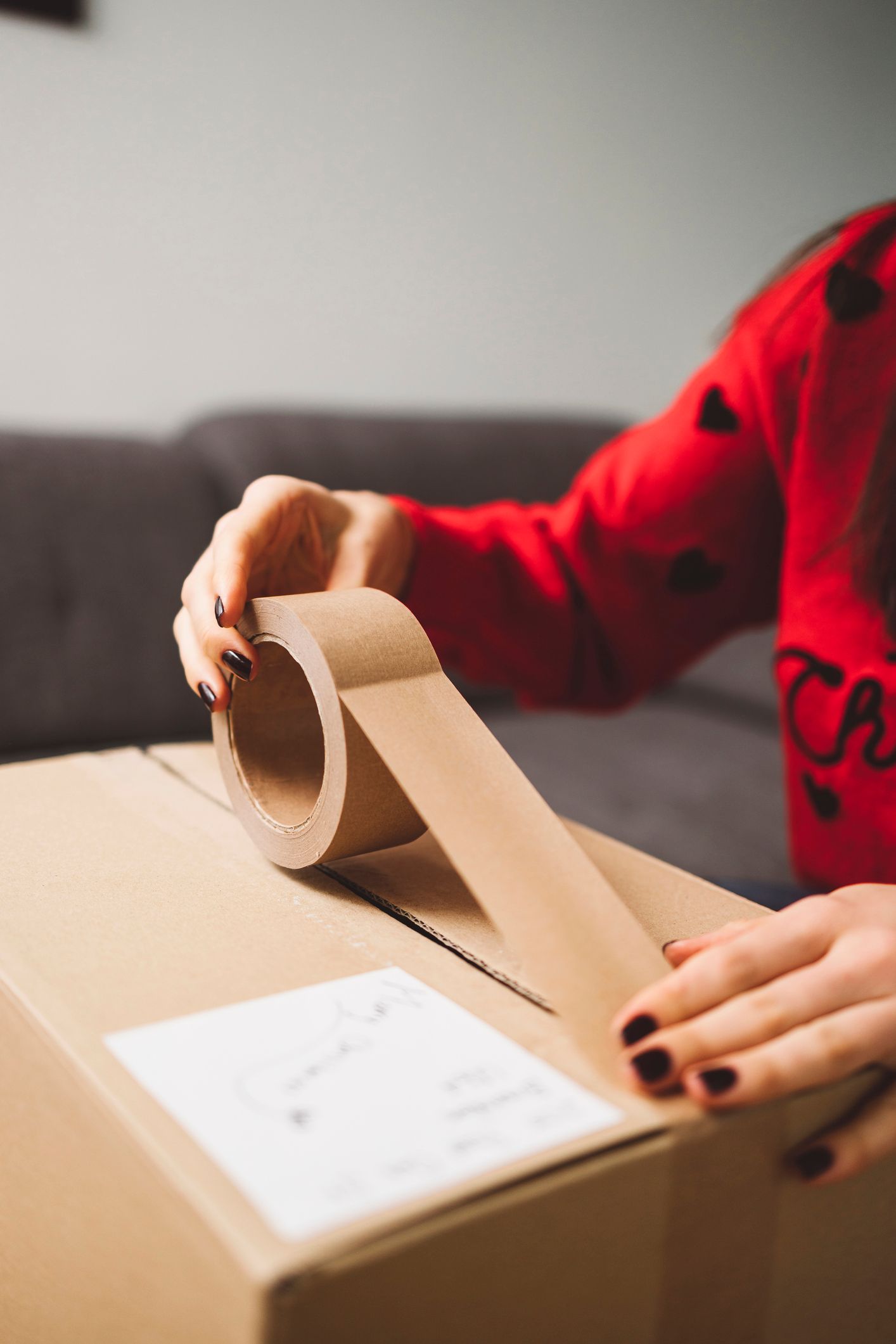 Person sealing a cardboard box with brown tape. They are wearing a red sweater and have dark nail polish.