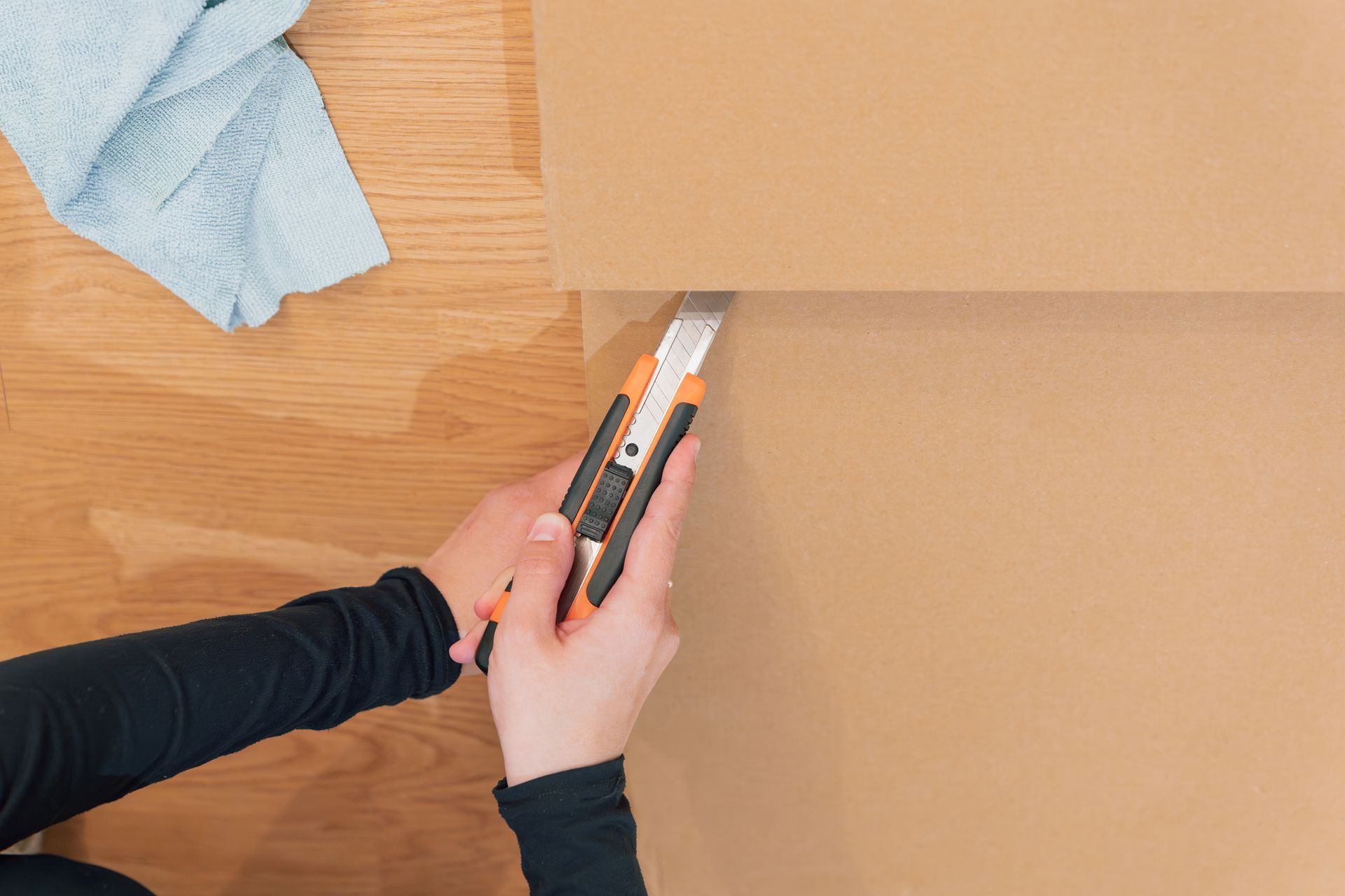 Person cutting cardboard with a utility knife on a wooden floor.