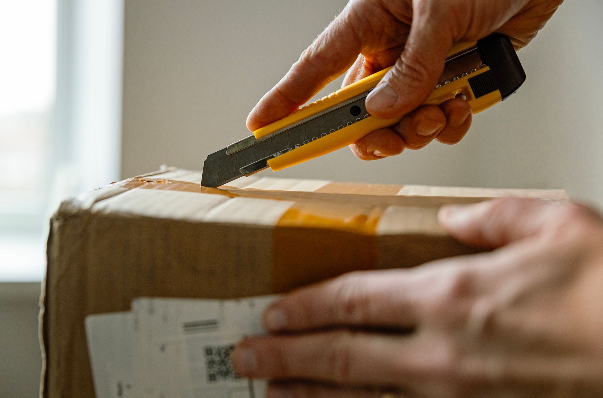 Person using a yellow box cutter to open a brown cardboard box, indoors.