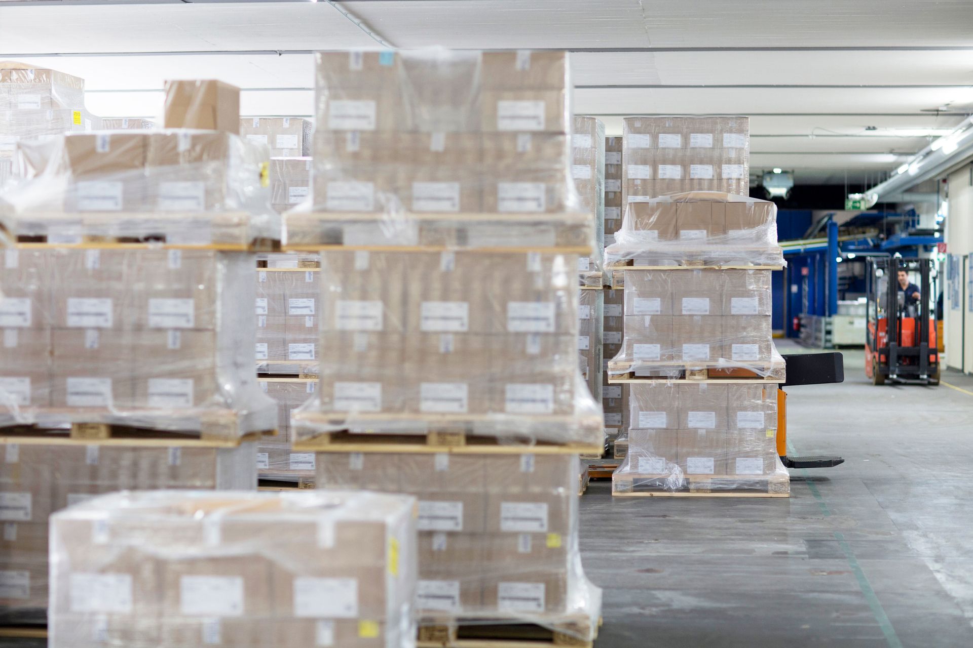 Pallets of wrapped boxes stacked in a warehouse; a forklift is in the background.