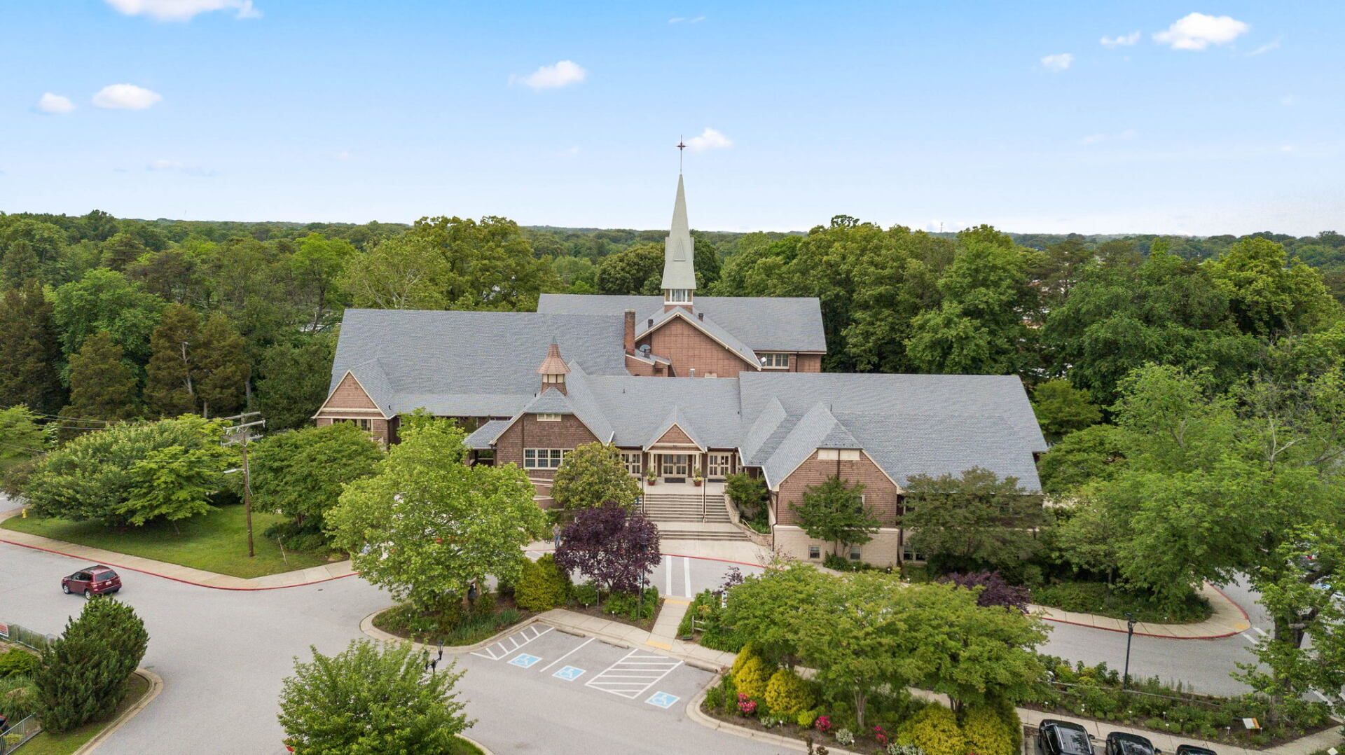 Aerial view of a brick church with a tall steeple surrounded by trees and a parking lot under a blue sky.