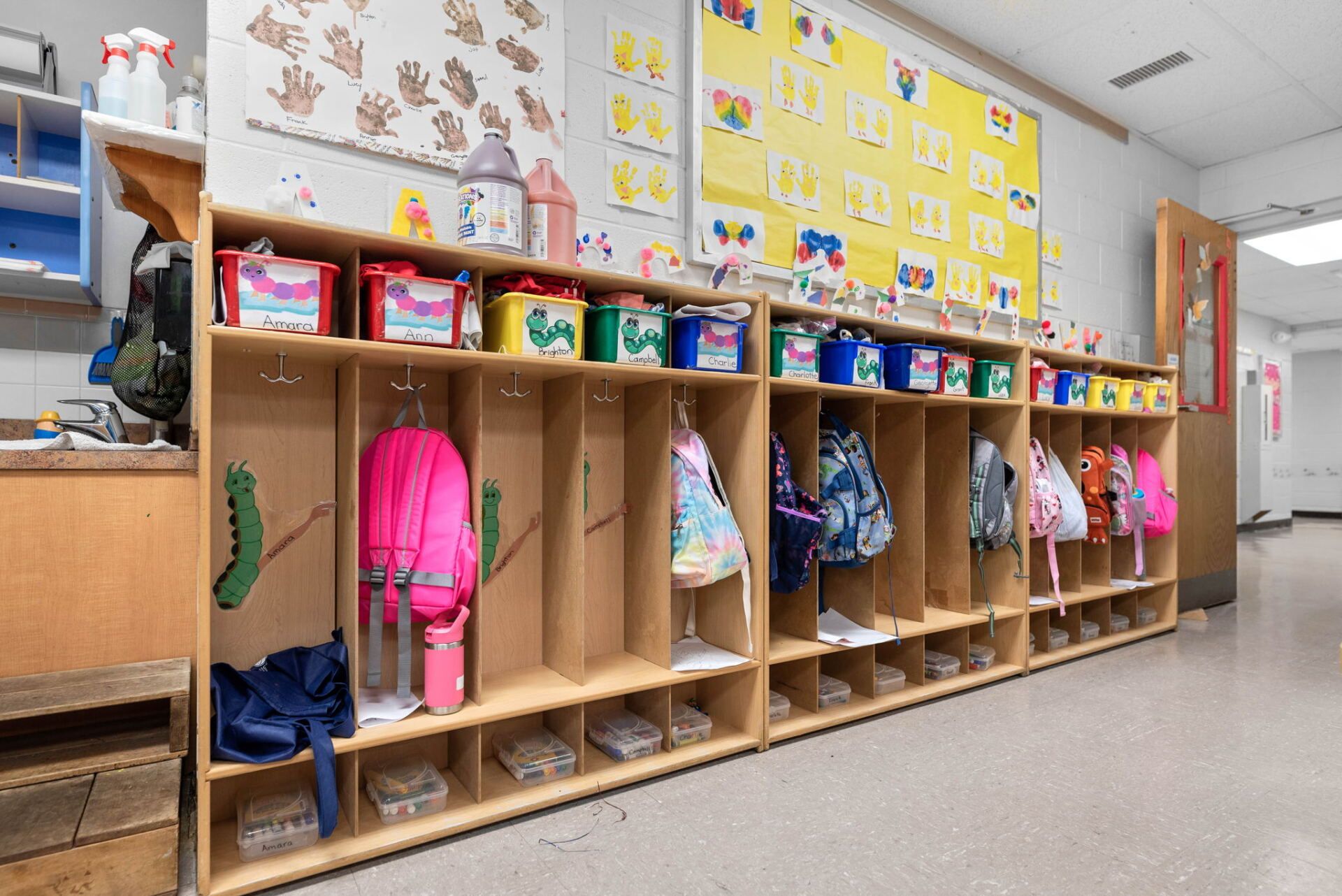 Row of wooden lockers in a preschool hallway, each with a hook and cubby, holding backpacks and supplies.