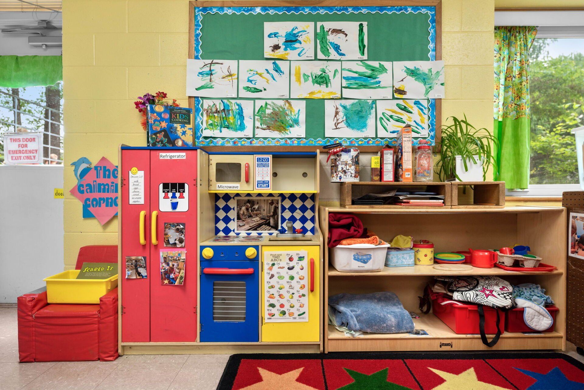 A colorful children's play area with a toy kitchen, artwork, and shelves filled with toys, set in a brightly lit room.