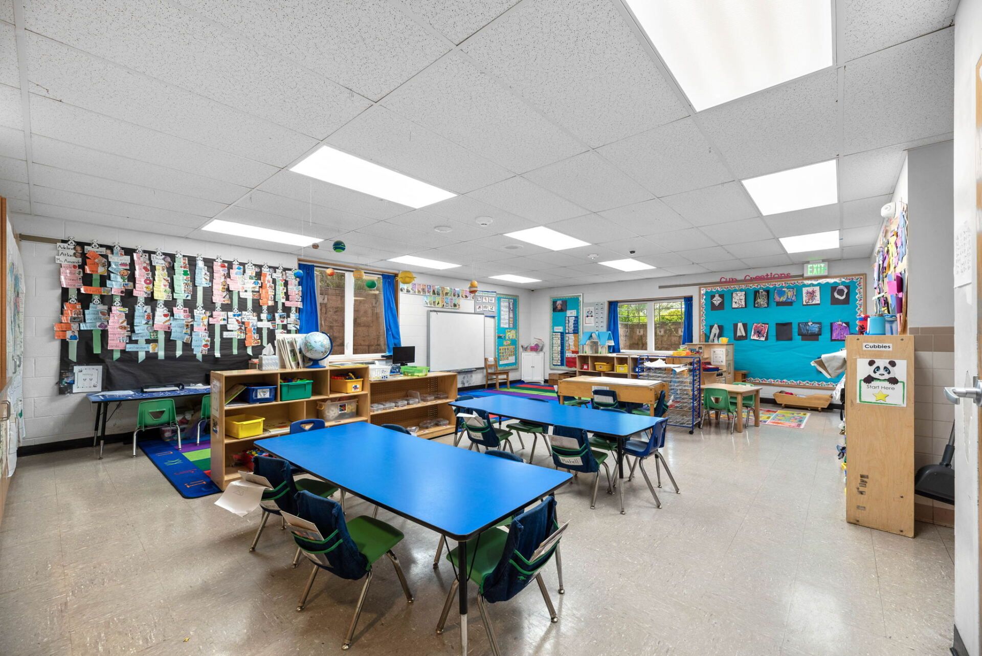 A brightly lit preschool classroom with blue tables, small chairs, and colorful artwork.