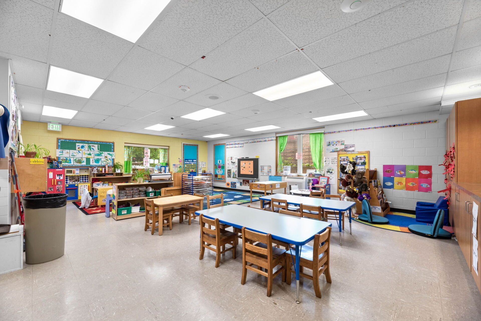 A brightly lit preschool classroom with small tables and chairs, bookshelves, and colorful decorations.