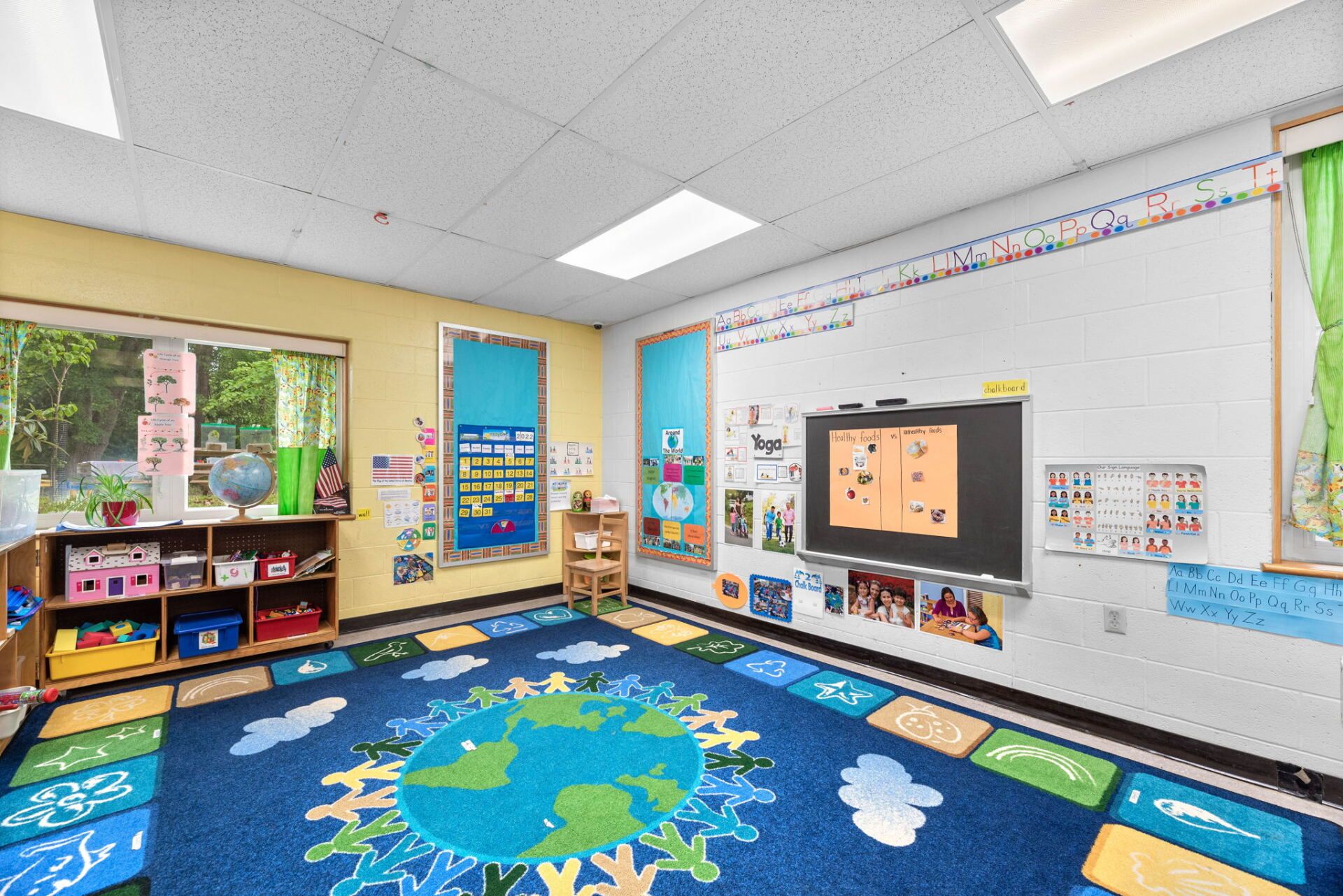 Classroom with a large Earth-themed rug. There's a whiteboard, colorful decorations, and a window overlooking greenery.