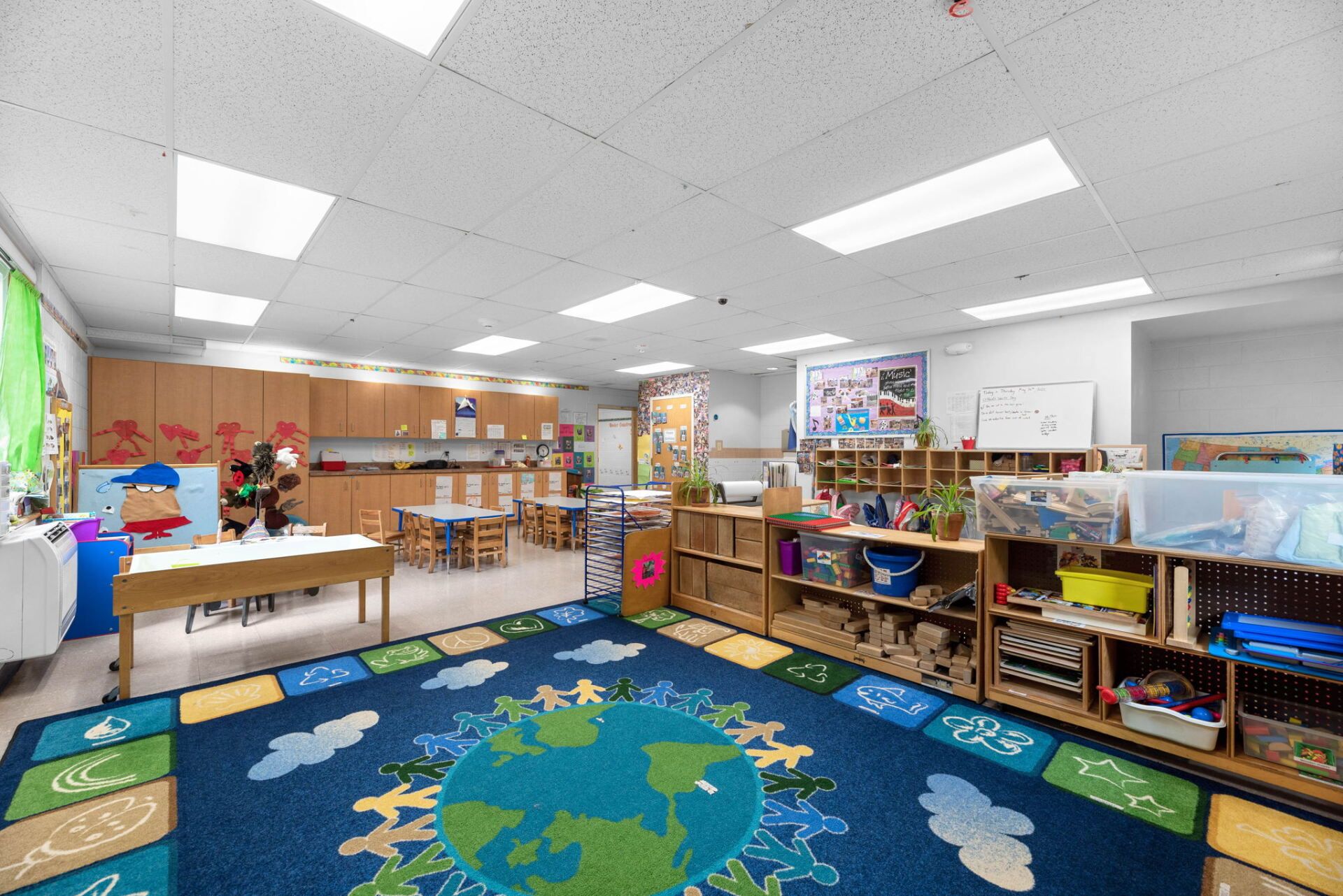 A colorful preschool classroom with a world map rug, wooden shelves with toys, and tables for children.