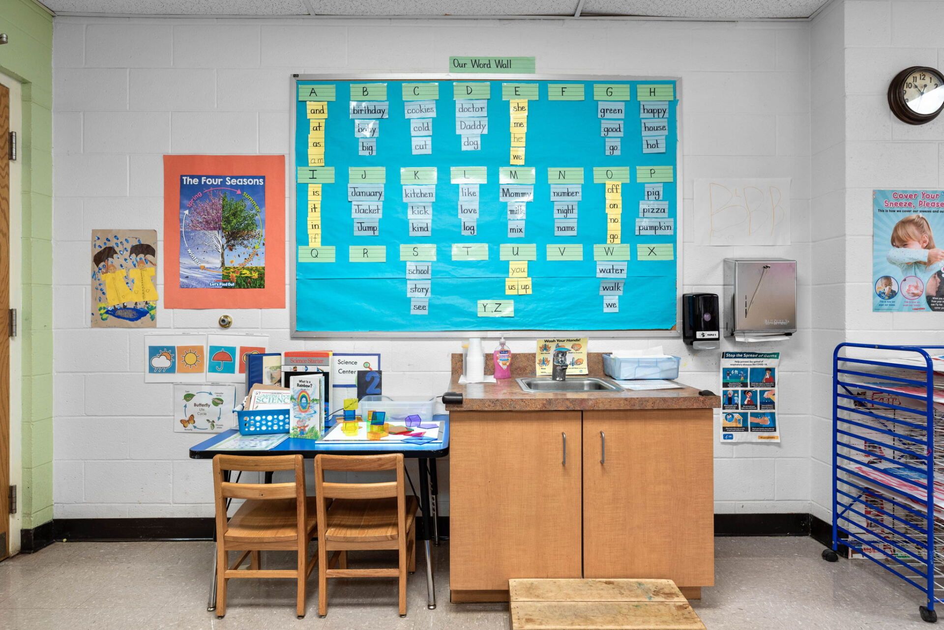 A classroom with a blue poster board, sink, small table, and various artwork on the white walls.