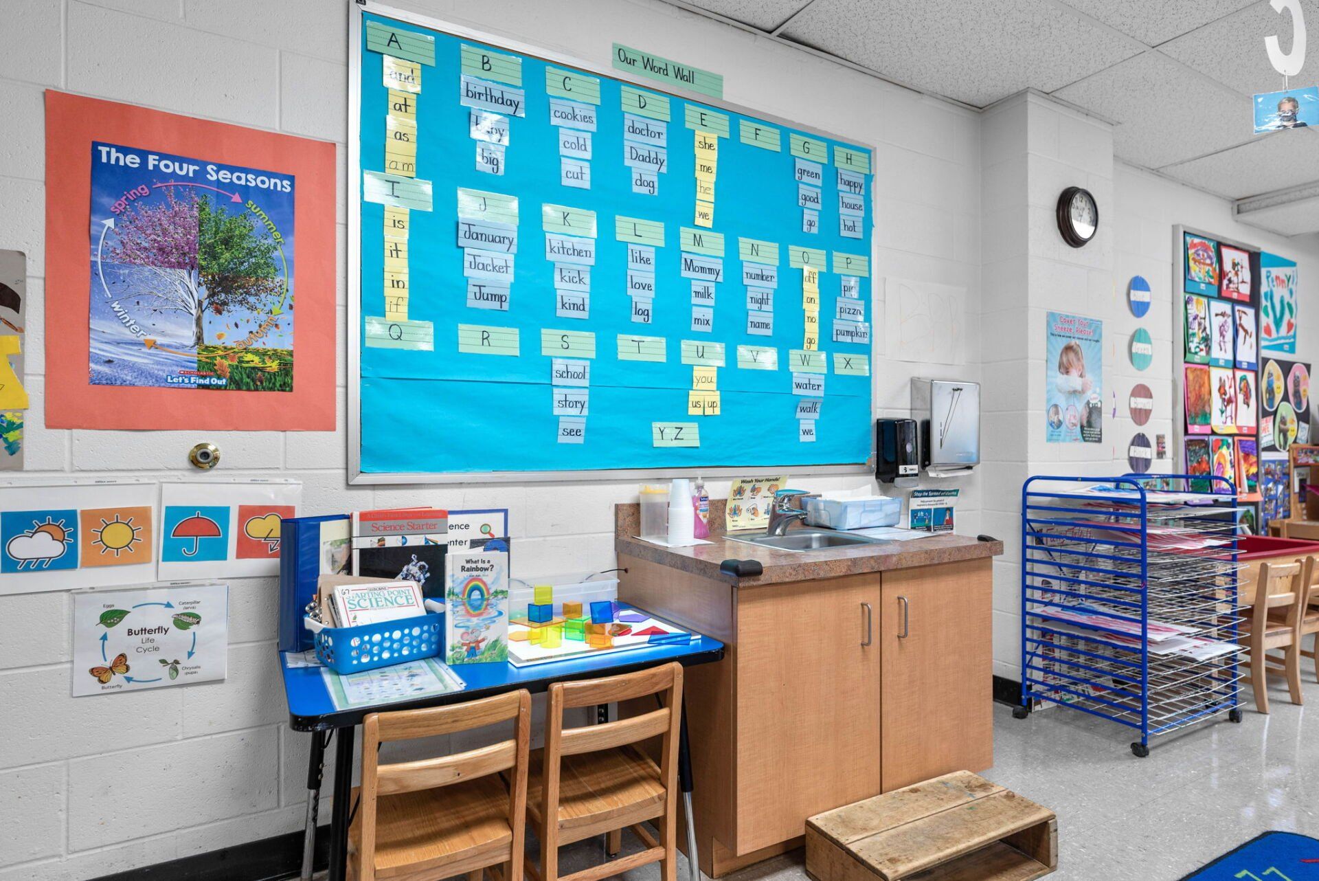 Classroom with a blue bulletin board, art, a desk with chairs, and a wooden cabinet.