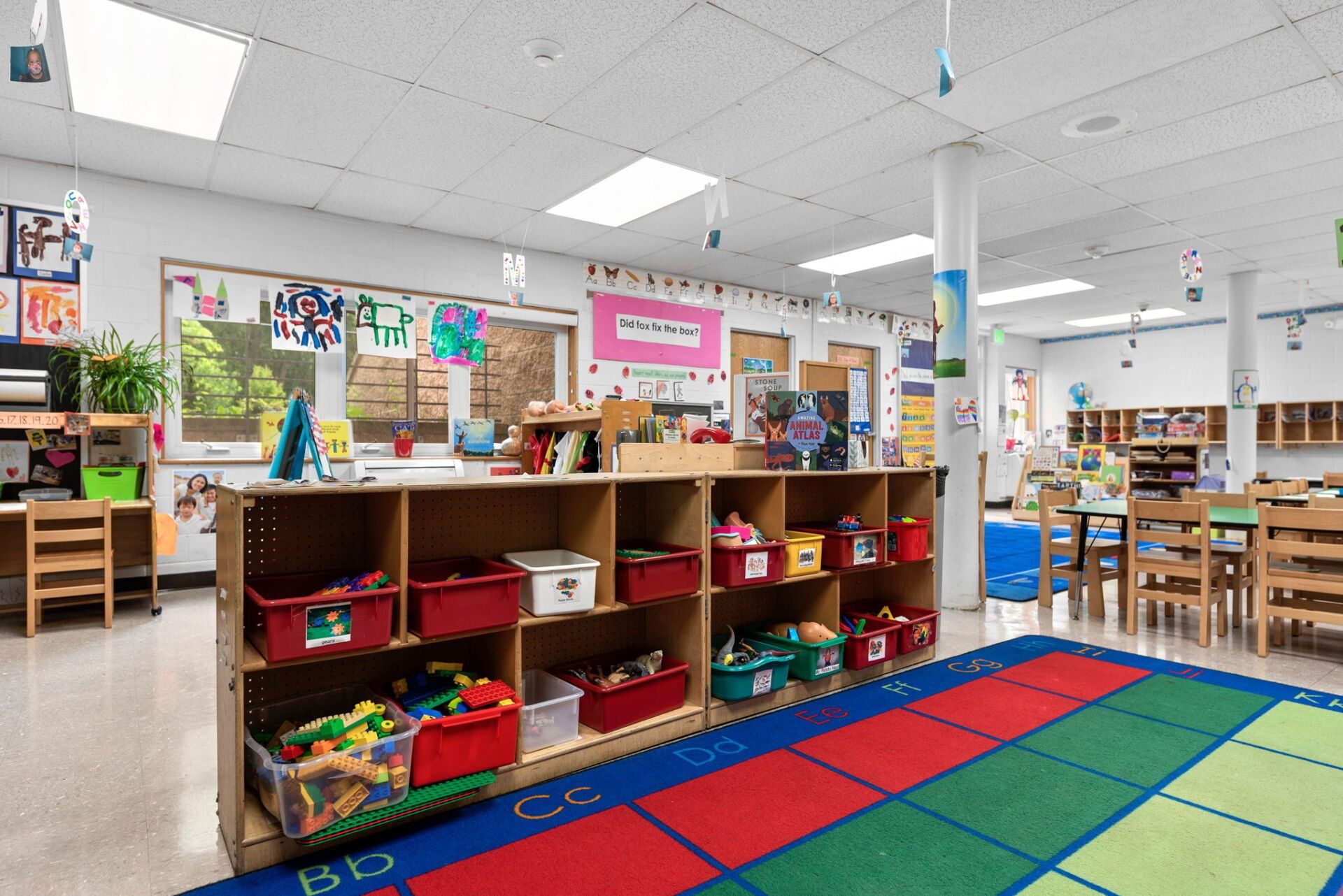 A brightly lit preschool classroom. Wooden shelves hold toys and supplies. A colorful ABC rug covers the floor.