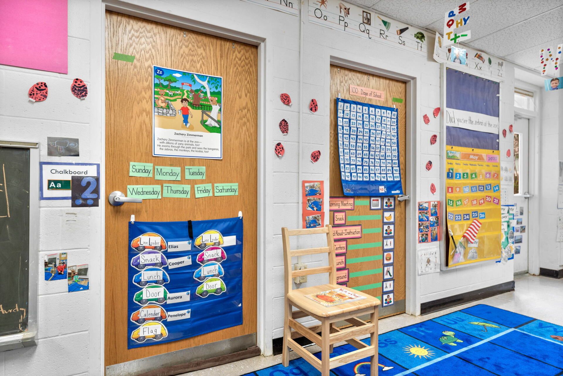 Hallway with two classroom doors decorated with educational materials. A small wooden chair sits in the middle. A blue rug is visible.