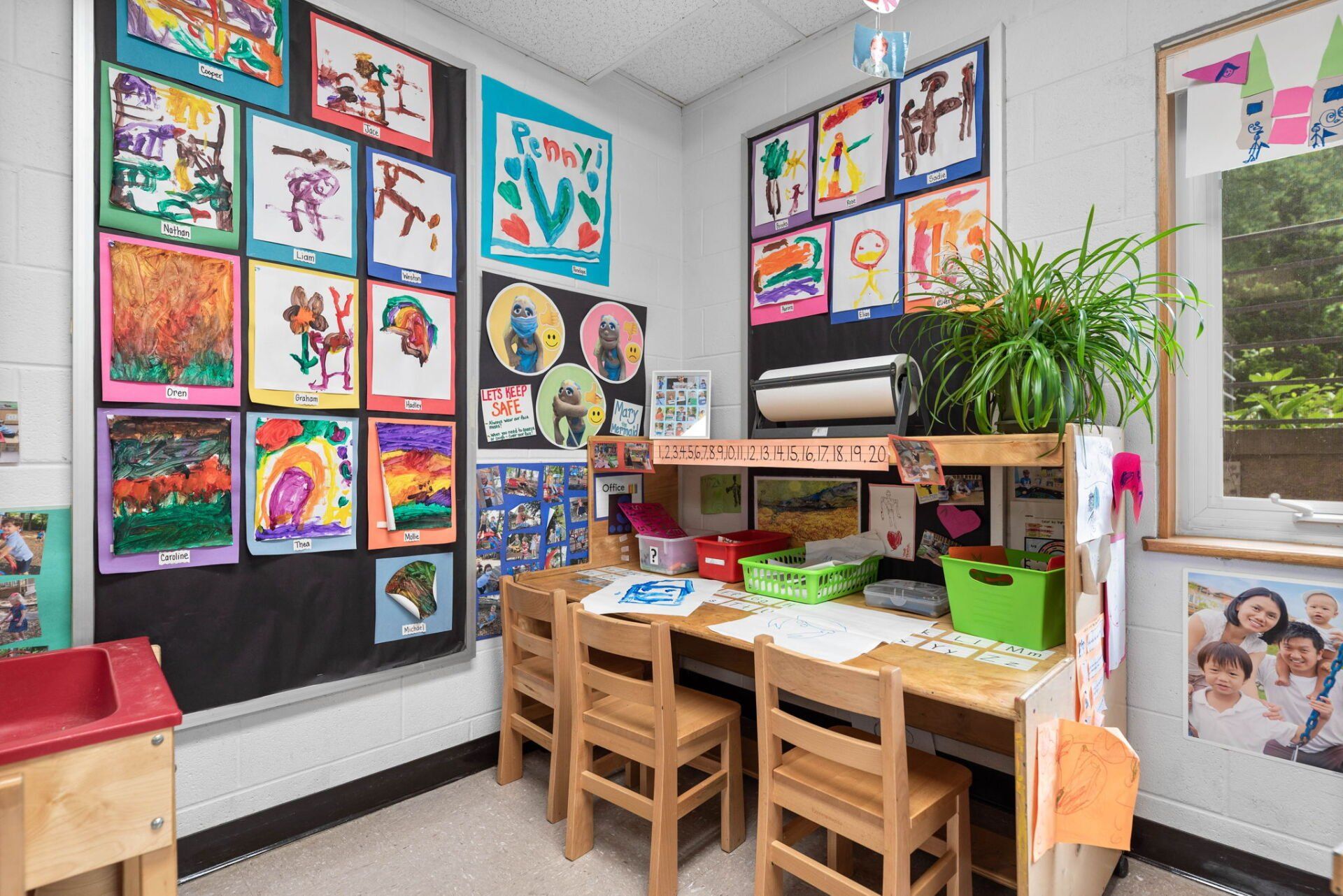 A brightly lit preschool art classroom with student artwork, wooden desks, and green plants.