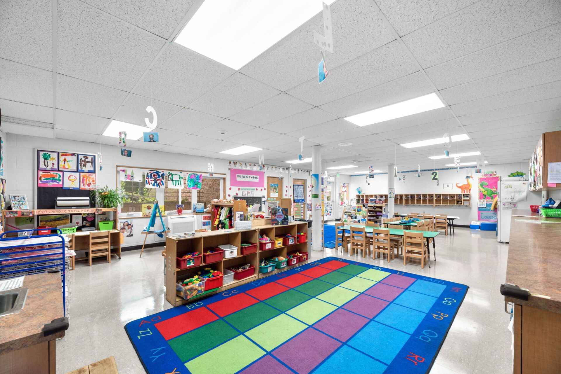 A bright, colorful preschool classroom with tables, shelves of toys, artwork on the walls, and a colorful rug.