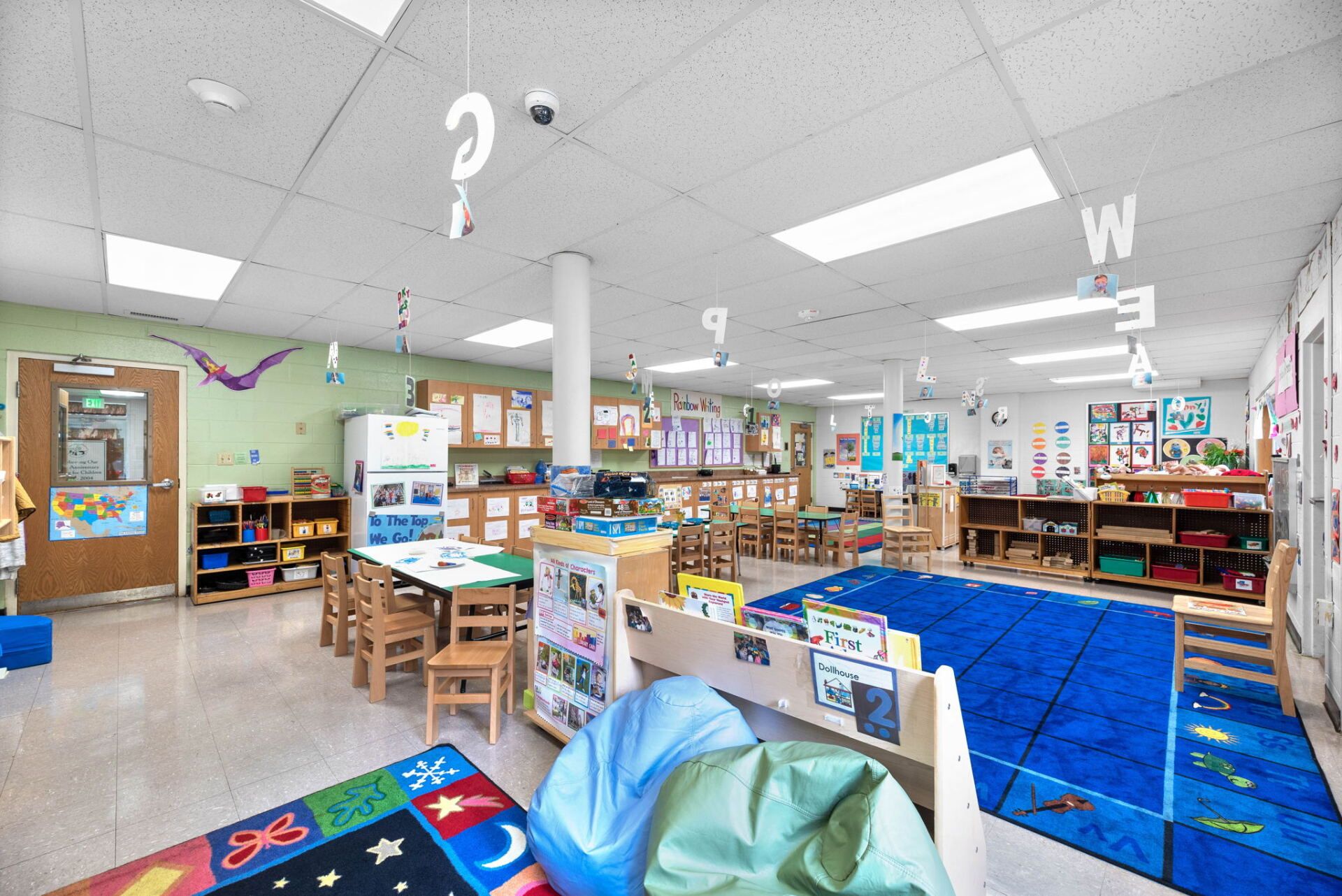 A bright, colorful classroom for young children, with a blue rug, wooden shelves, and various learning materials.