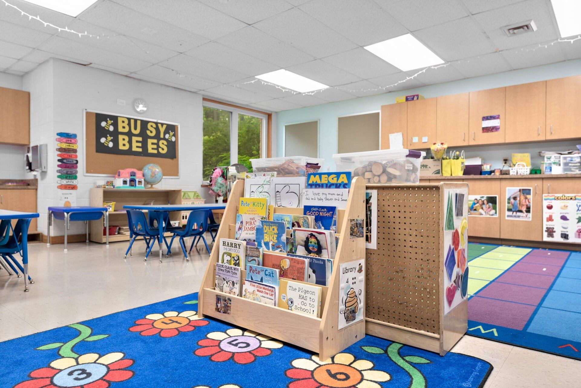 Classroom interior: Bookshelf with children's books, tables, and a colorful rug.