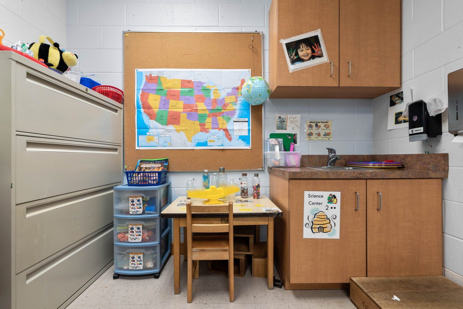 A classroom corner with a small table, sink, storage cabinets, and a map of the United States.
