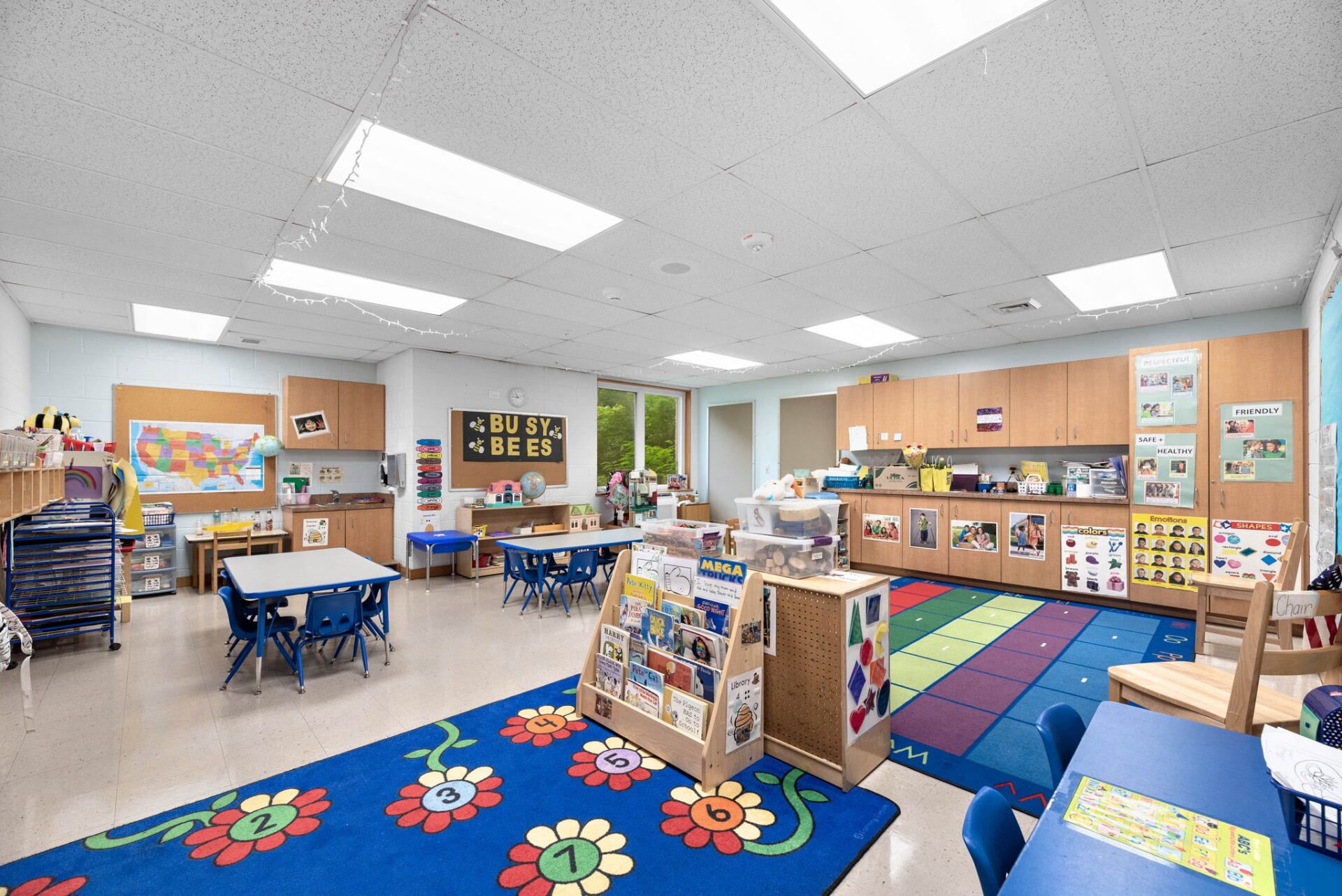 Bright, airy classroom with blue tables, colorful rugs, and wooden storage units. Children's toys and learning materials are visible.