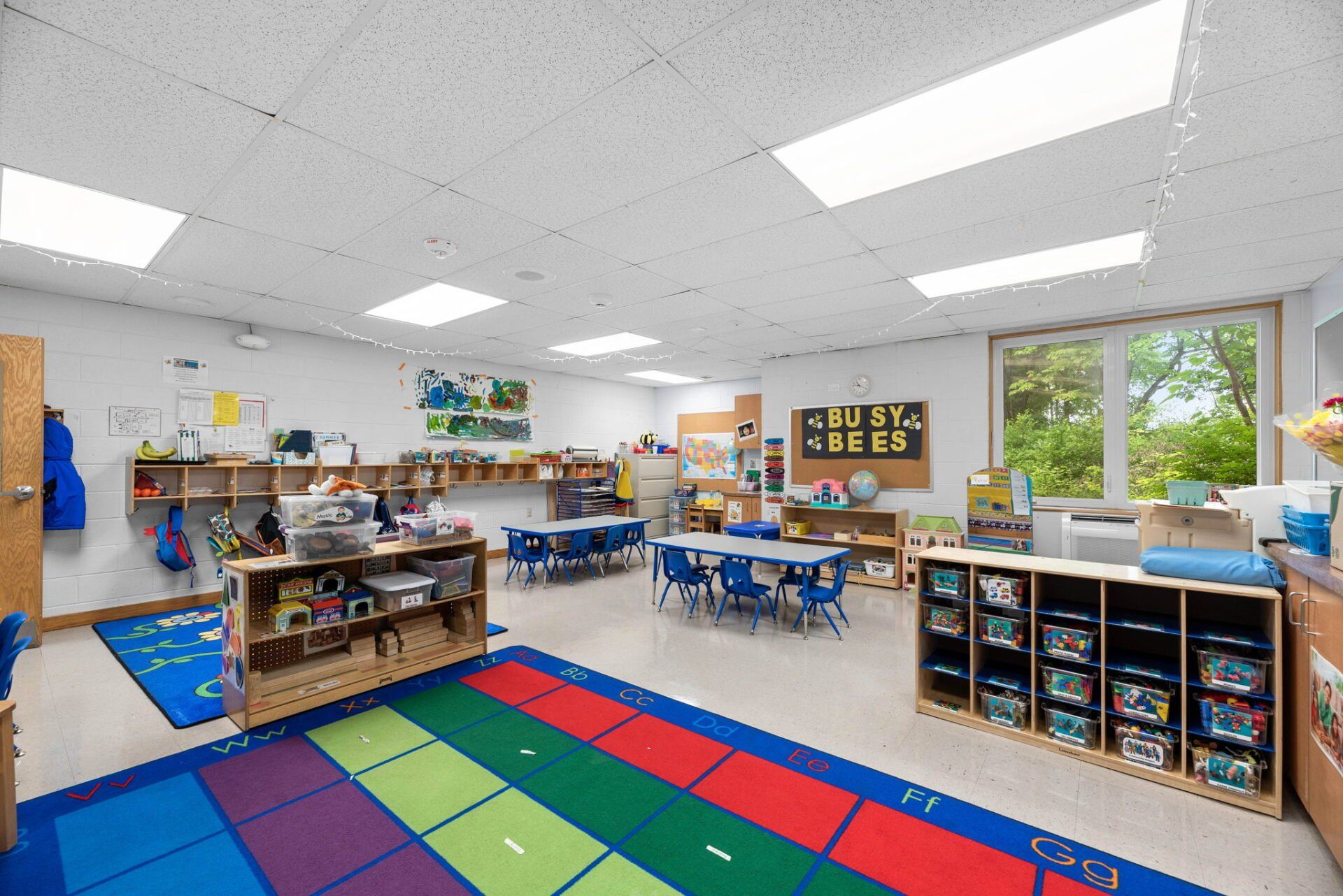 A well-lit preschool classroom with colorful rugs, small tables, and shelves filled with supplies. A large window offers a view of trees outside.