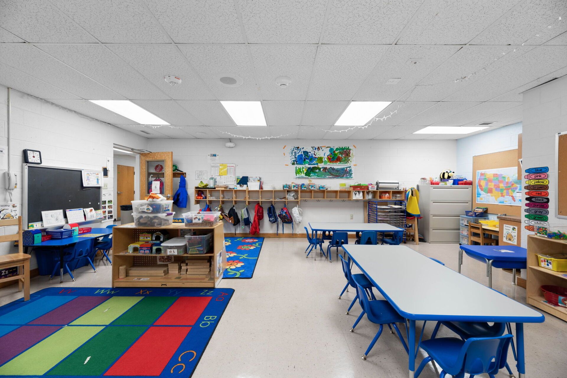 Empty preschool classroom with blue tables and chairs, a play rug, and various storage and art supplies.
