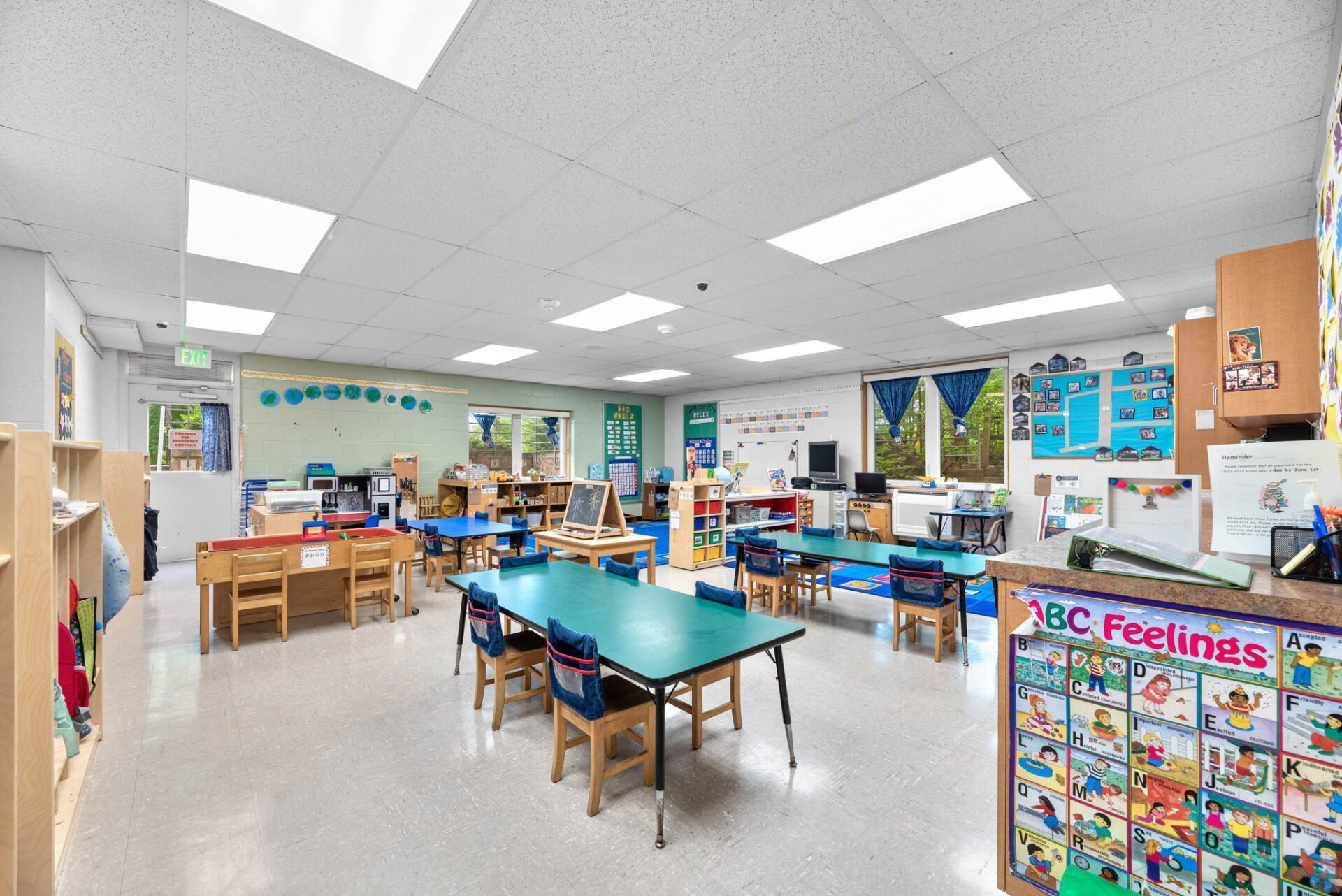 Brightly lit preschool classroom with colorful tables and chairs, bulletin boards, and shelves filled with books and toys.
