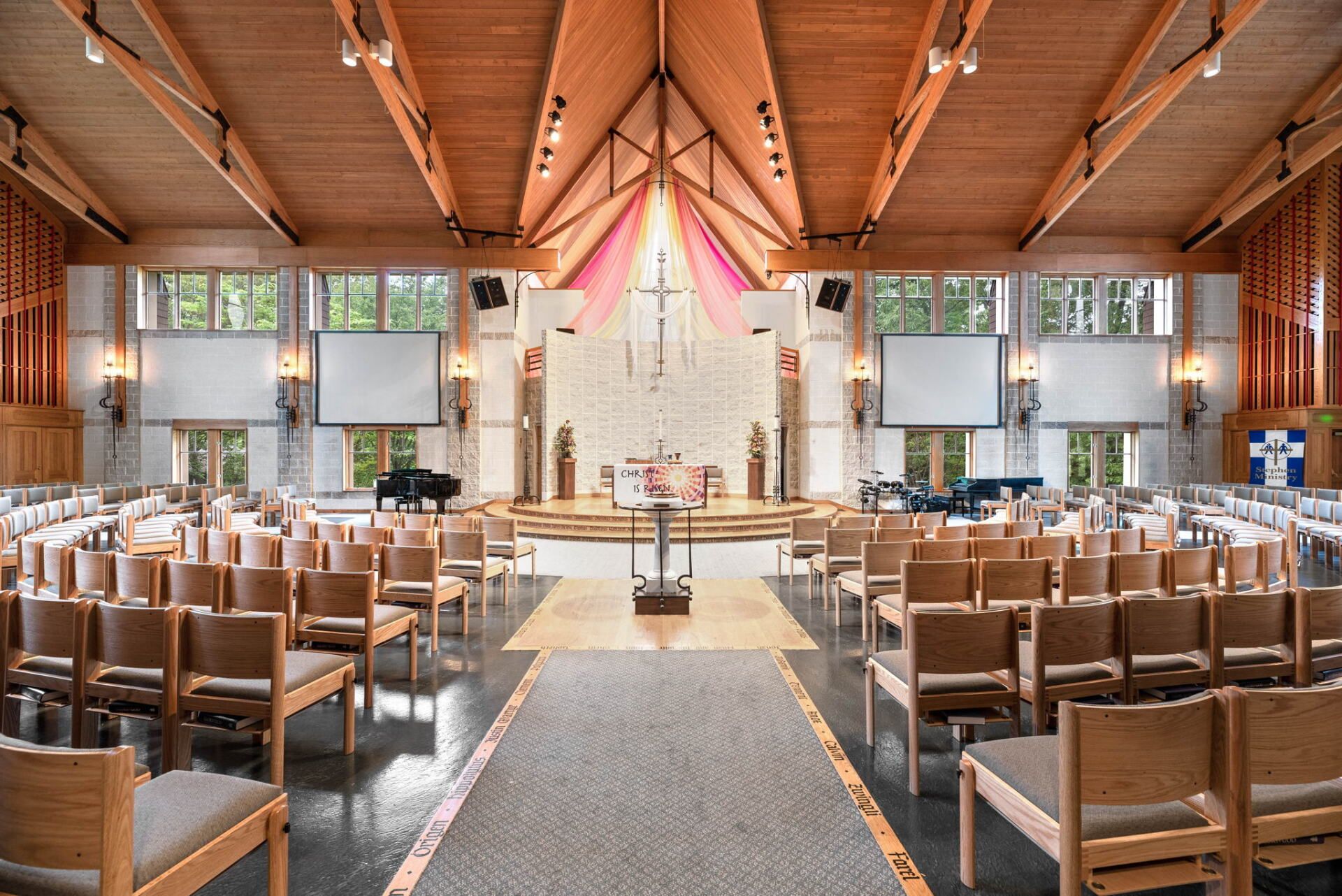 Interior of a modern church with wooden beams, rows of chairs, and a stage with a cross and altar. Sunlight streams in.