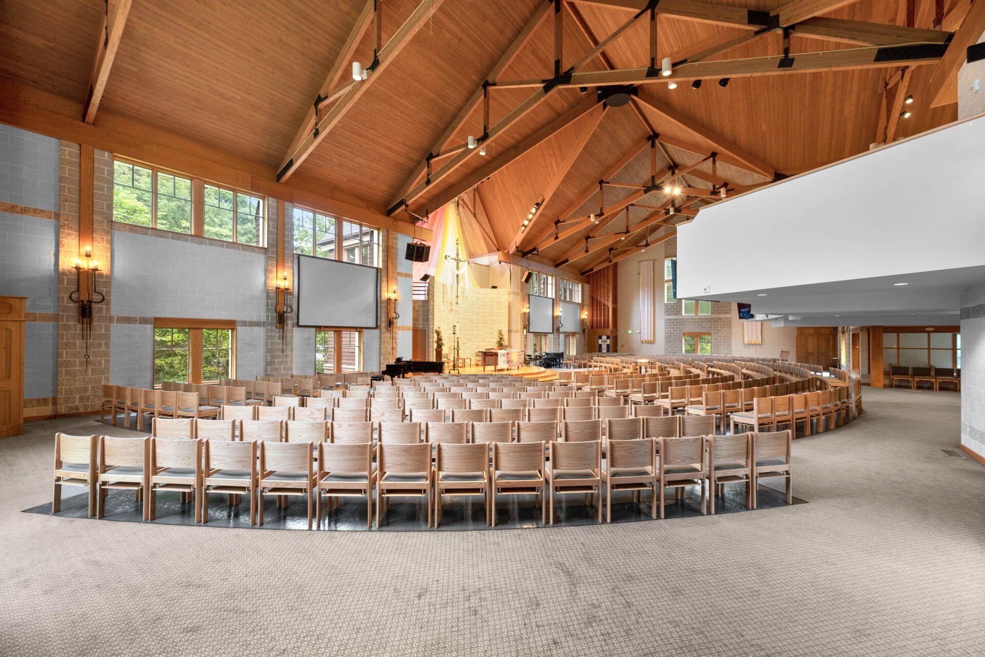 Interior of a spacious auditorium with a wooden vaulted ceiling, rows of chairs arranged in semi-circles, and large windows.