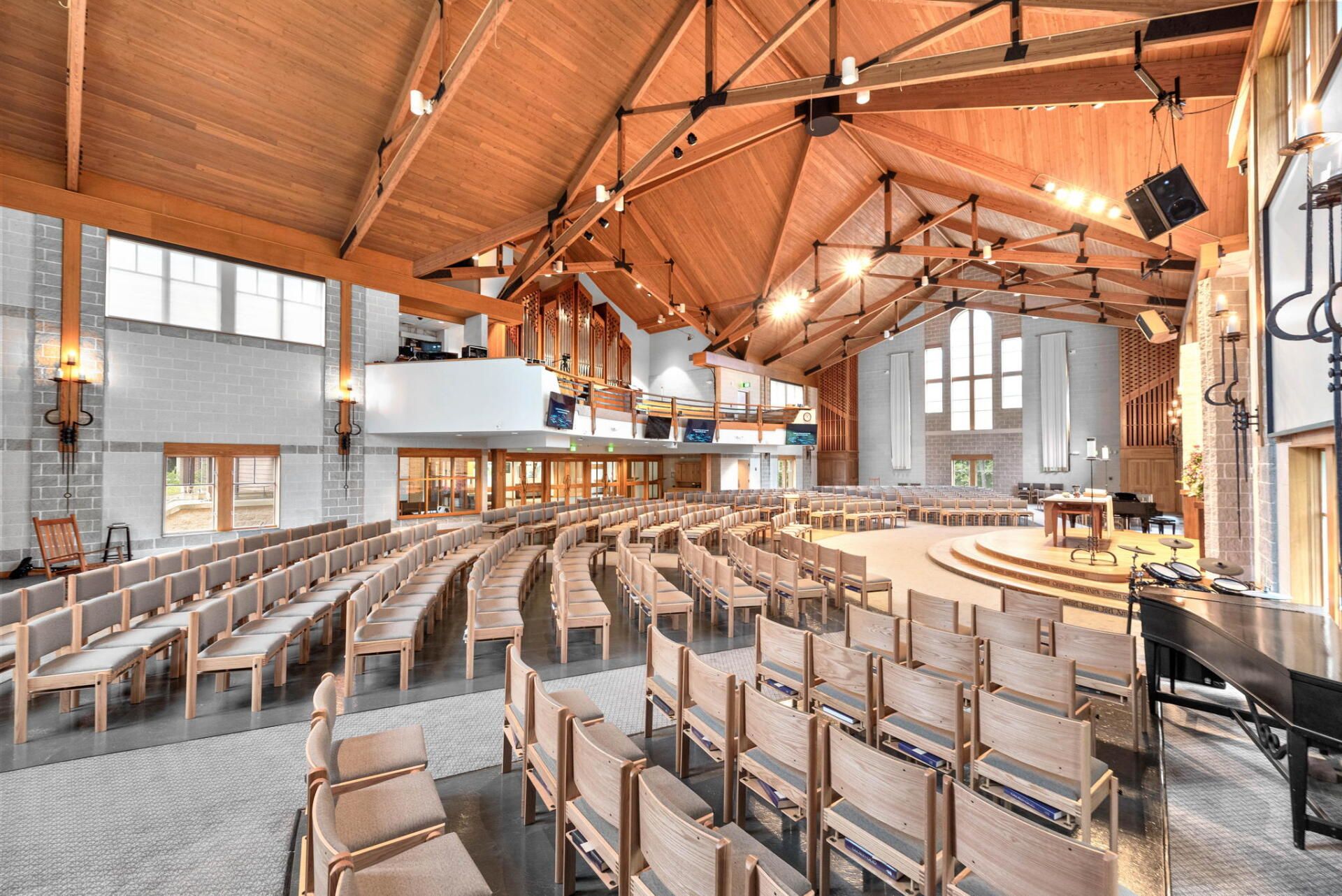Interior of a light-filled church sanctuary with curved rows of wooden pews facing a raised platform. Wooden beams and a large window are visible.
