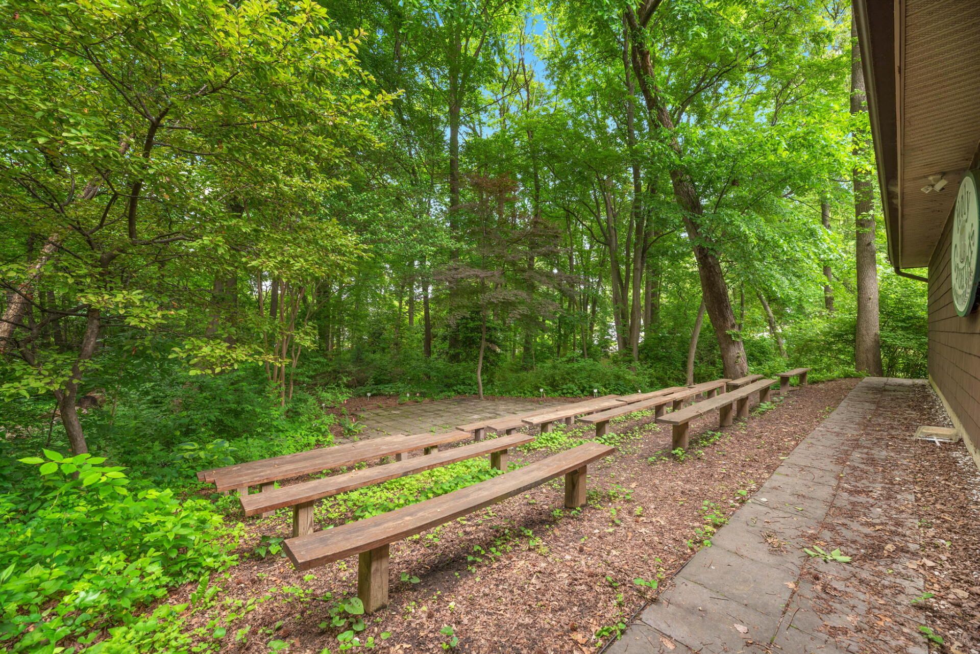 Wooden benches face a wooded area. A brick path runs alongside a building's exterior. Green foliage surrounds the scene.