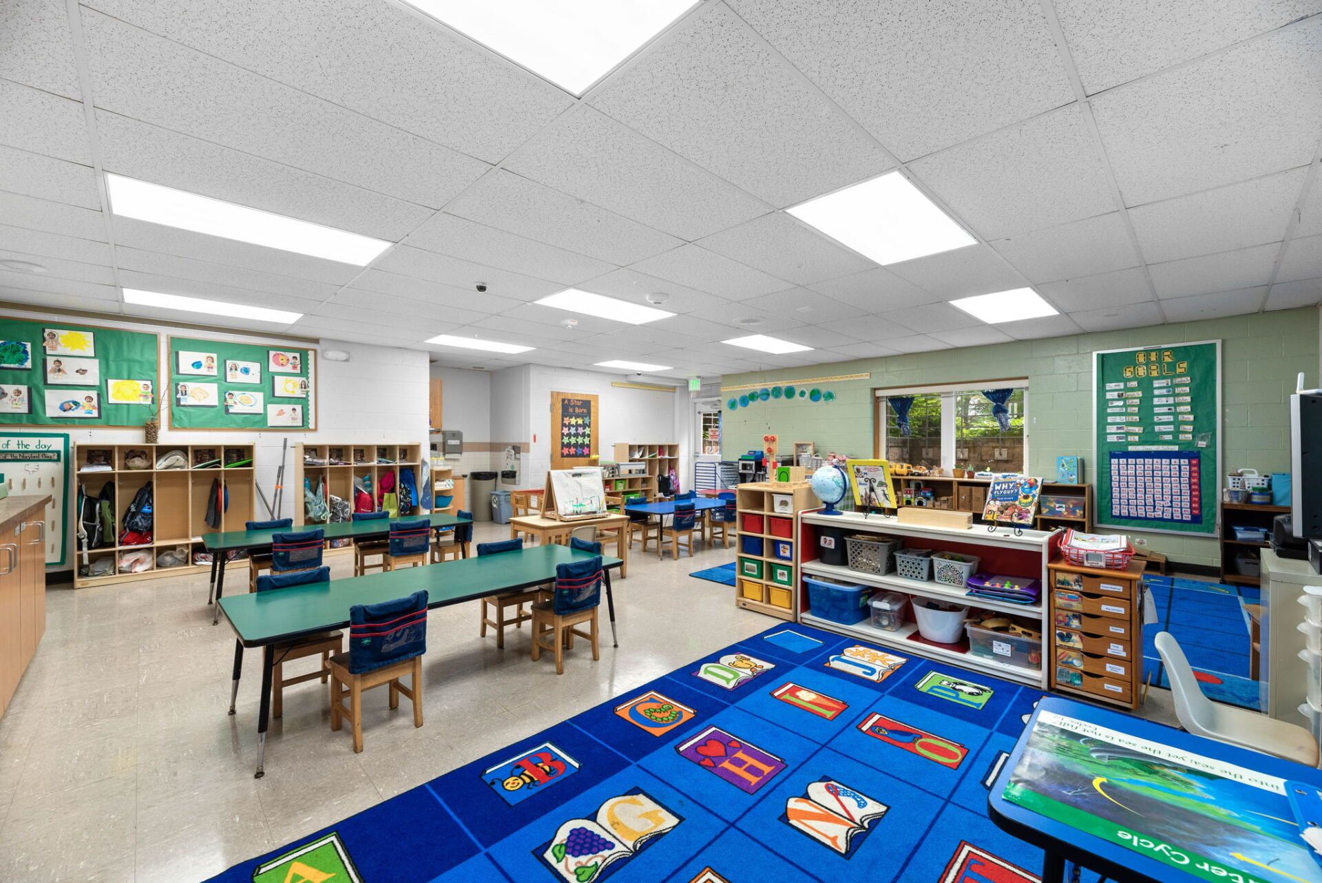 Empty preschool classroom with blue and green tables, a colorful rug, and shelves filled with books and toys.