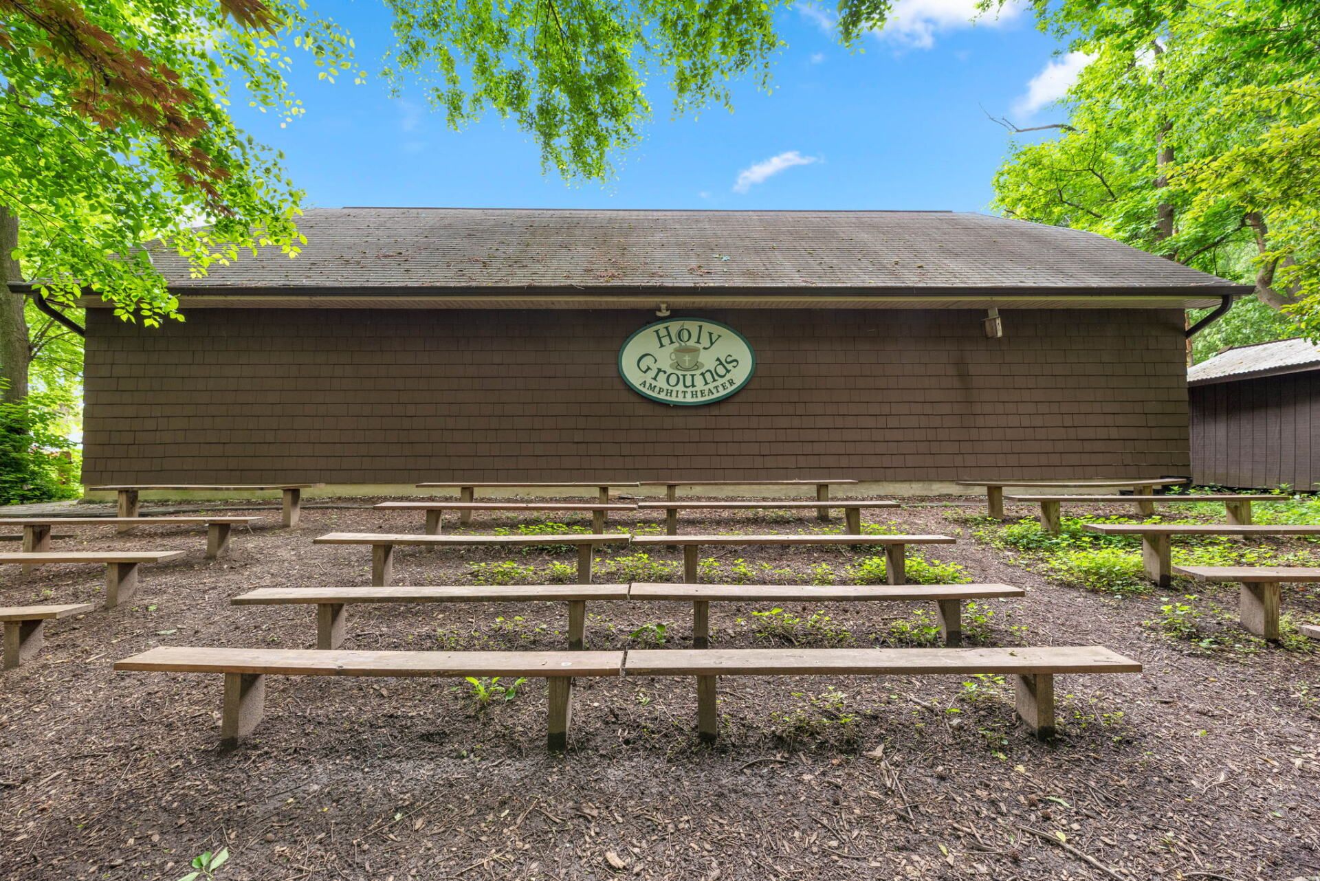 An outdoor theater with tiered wooden benches. Brown building with a sign stands at the back, surrounded by trees.