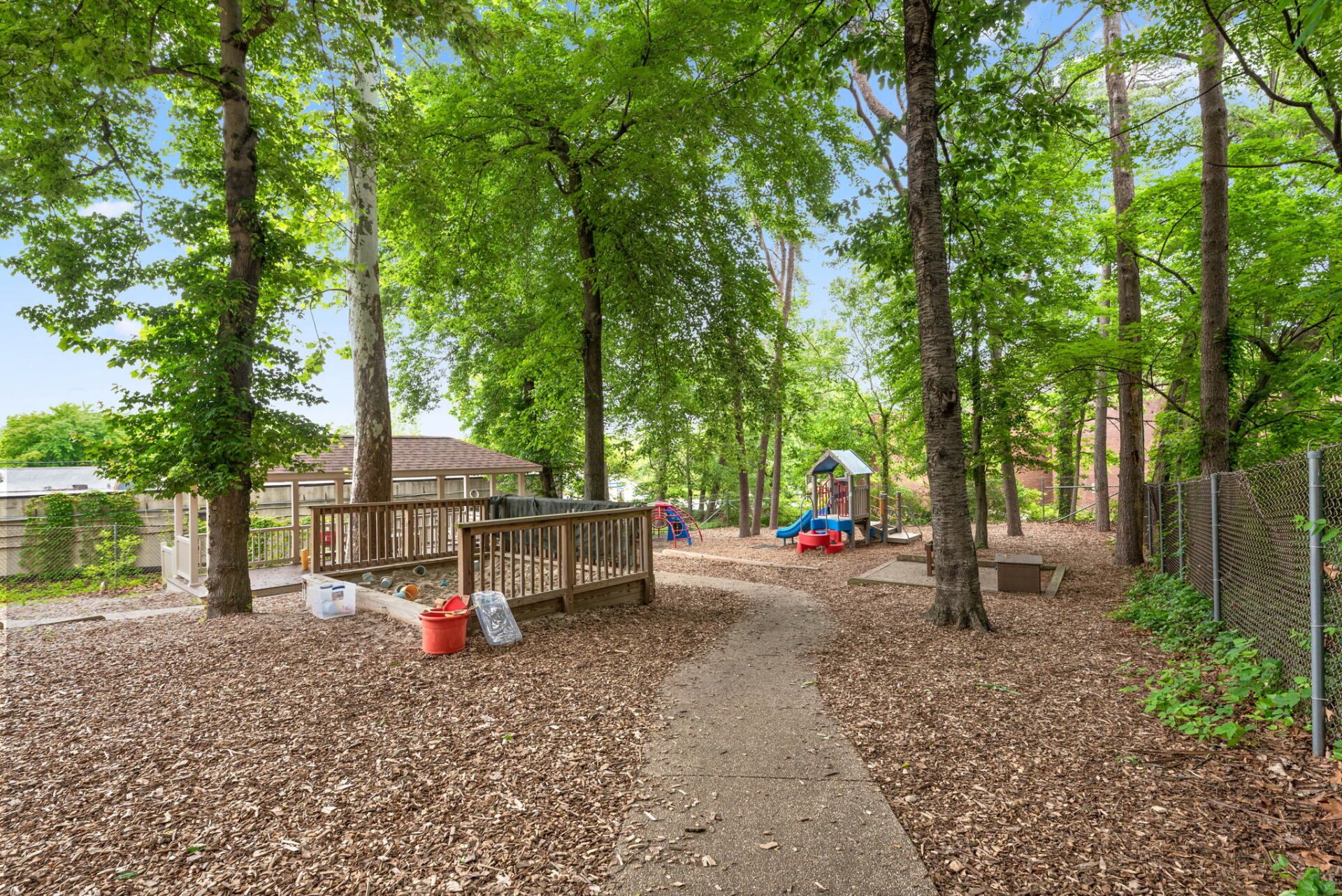 A park with a playground, wooden deck, trees, and path covered in leaves. Children's toys are visible.