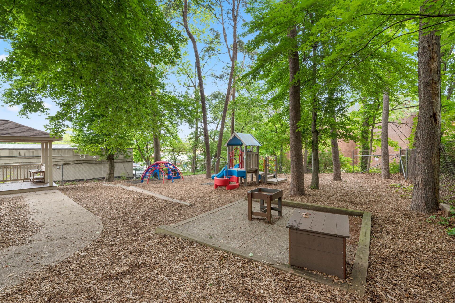 Playground in a wooded area, featuring play structures, a concrete patio, and a picnic table. Covered in fallen leaves.